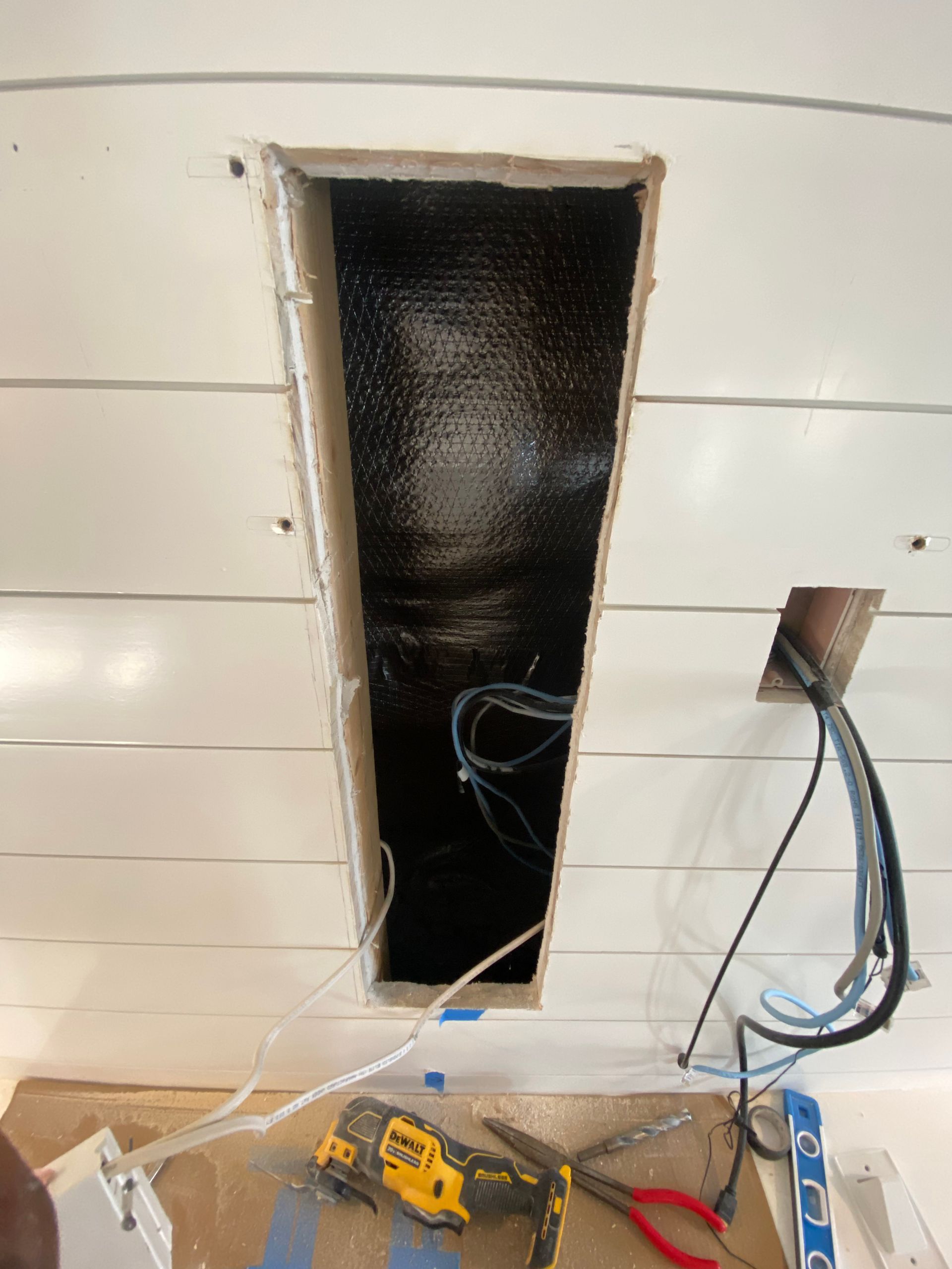 A rectangular opening in a white shiplap wall reveals interior wiring, with tools and wires visible on the floor below.