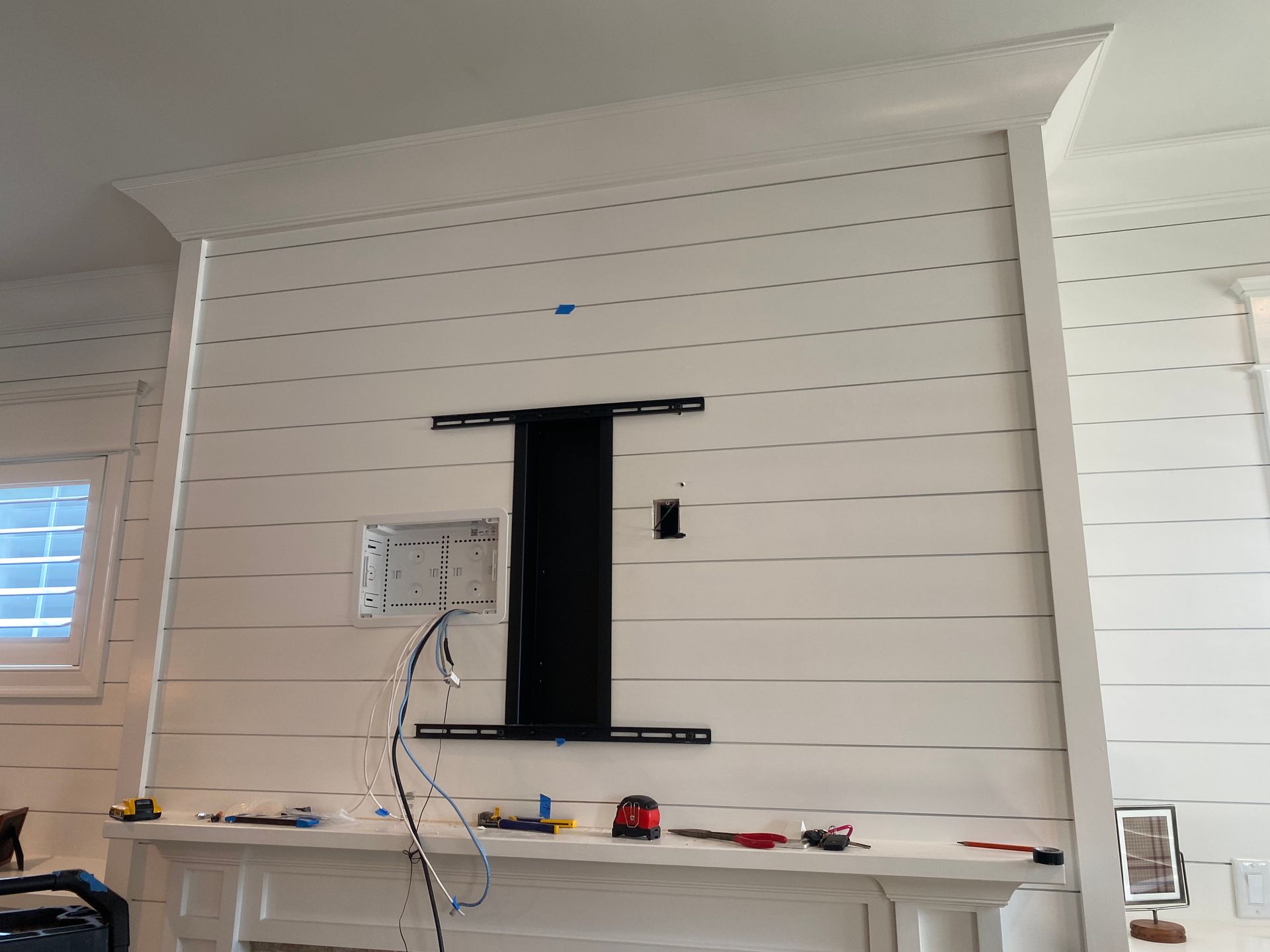 A wall with white shiplap, a fireplace mantle, and a mounted TV bracket with visible electrical wires and a wall outlet.