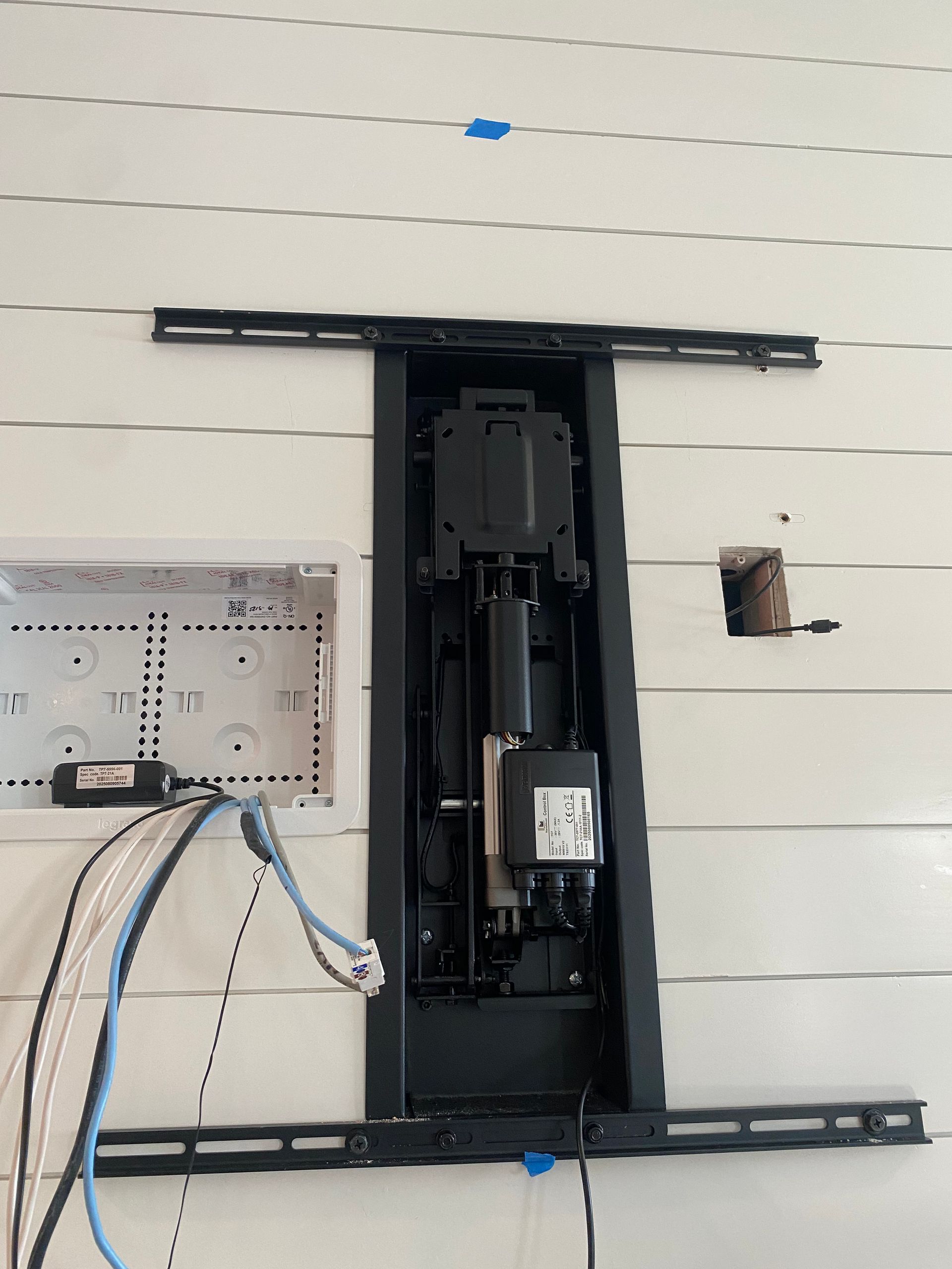 A motorized TV wall mount installed on a white paneled wall with exposed wiring and an open junction box to the left.