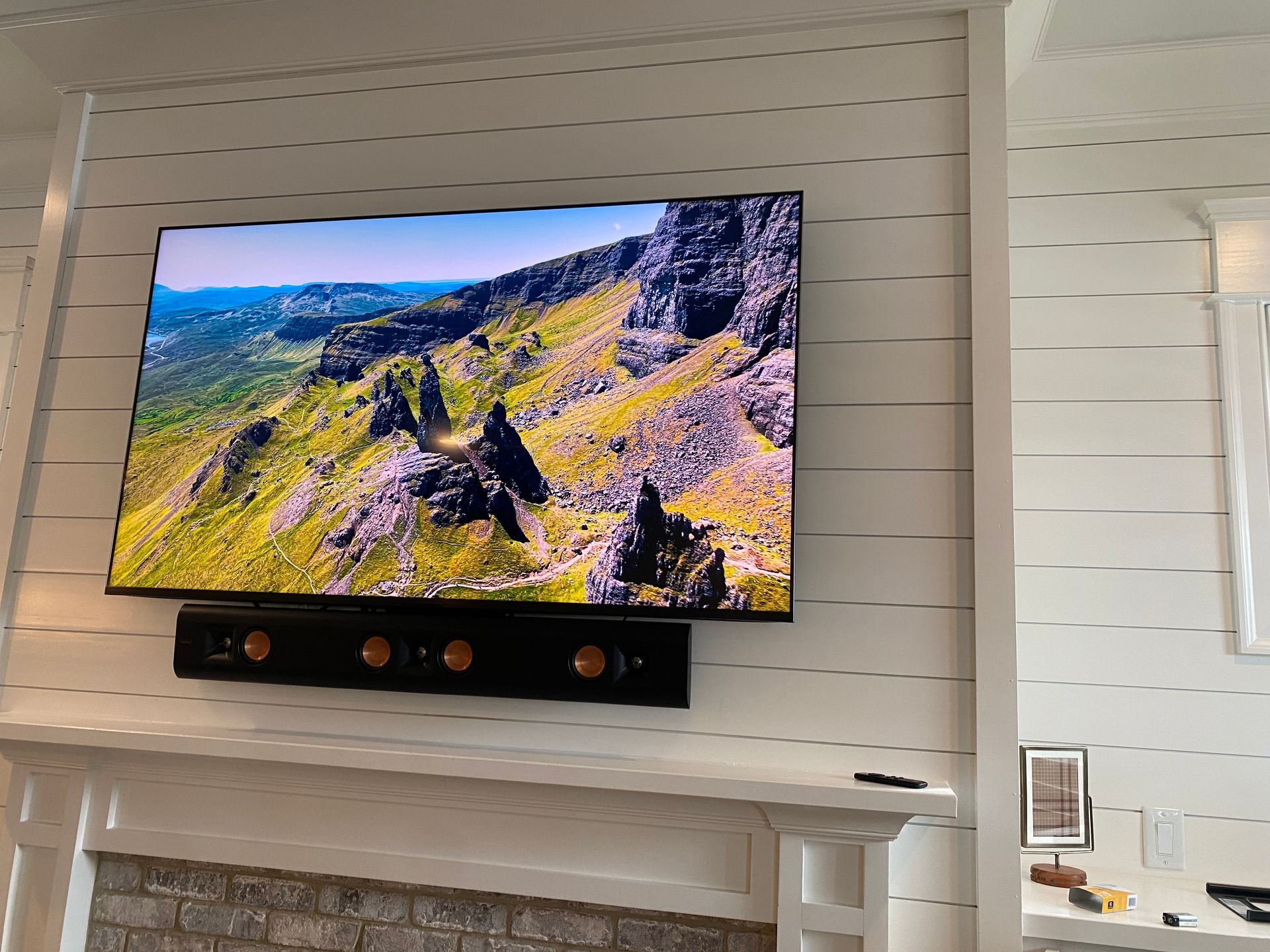 A large television showing a mountain landscape mounted on a white shiplap wall above a brick fireplace and soundbar.