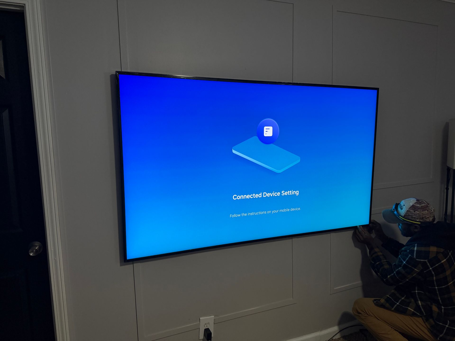 A person stands on the right, connecting a television screen to the wall, which displays a device setup blue screen.