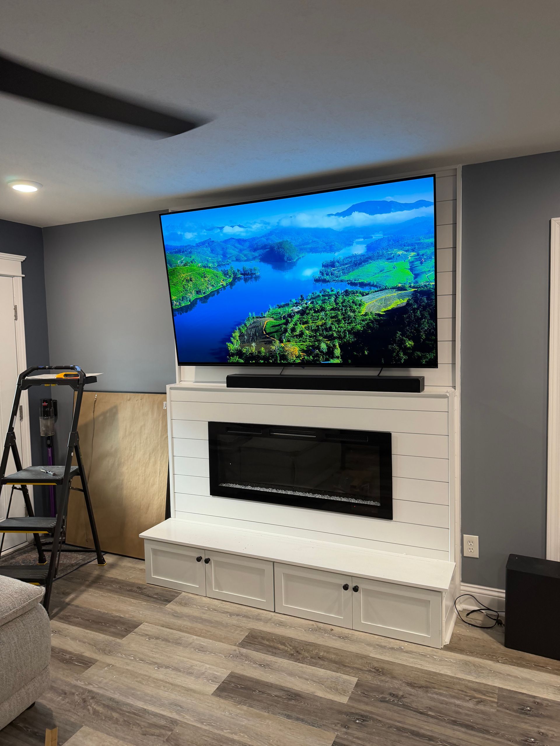 A wall-mounted TV and electric fireplace are built into a white shiplap feature wall in a room with gray walls.