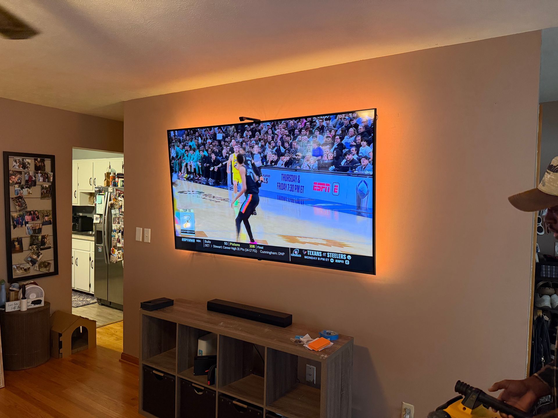 A wall-mounted TV showing a basketball game, illuminated by backlighting, sits above a wooden storage console in a home.