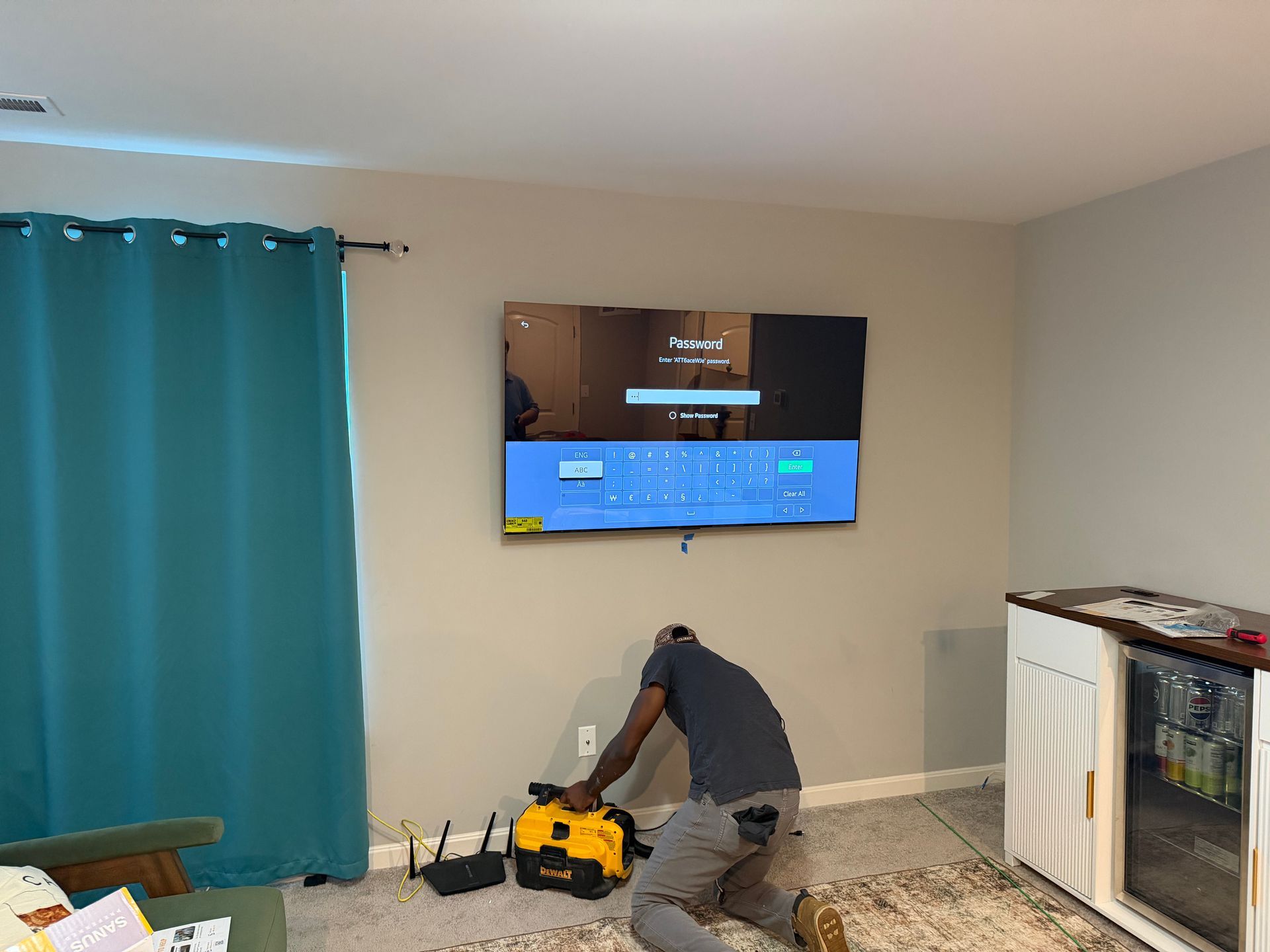 A worker kneels on a rug while installing a wall-mounted TV with a blue screen next to teal curtains and a beverage fridge.