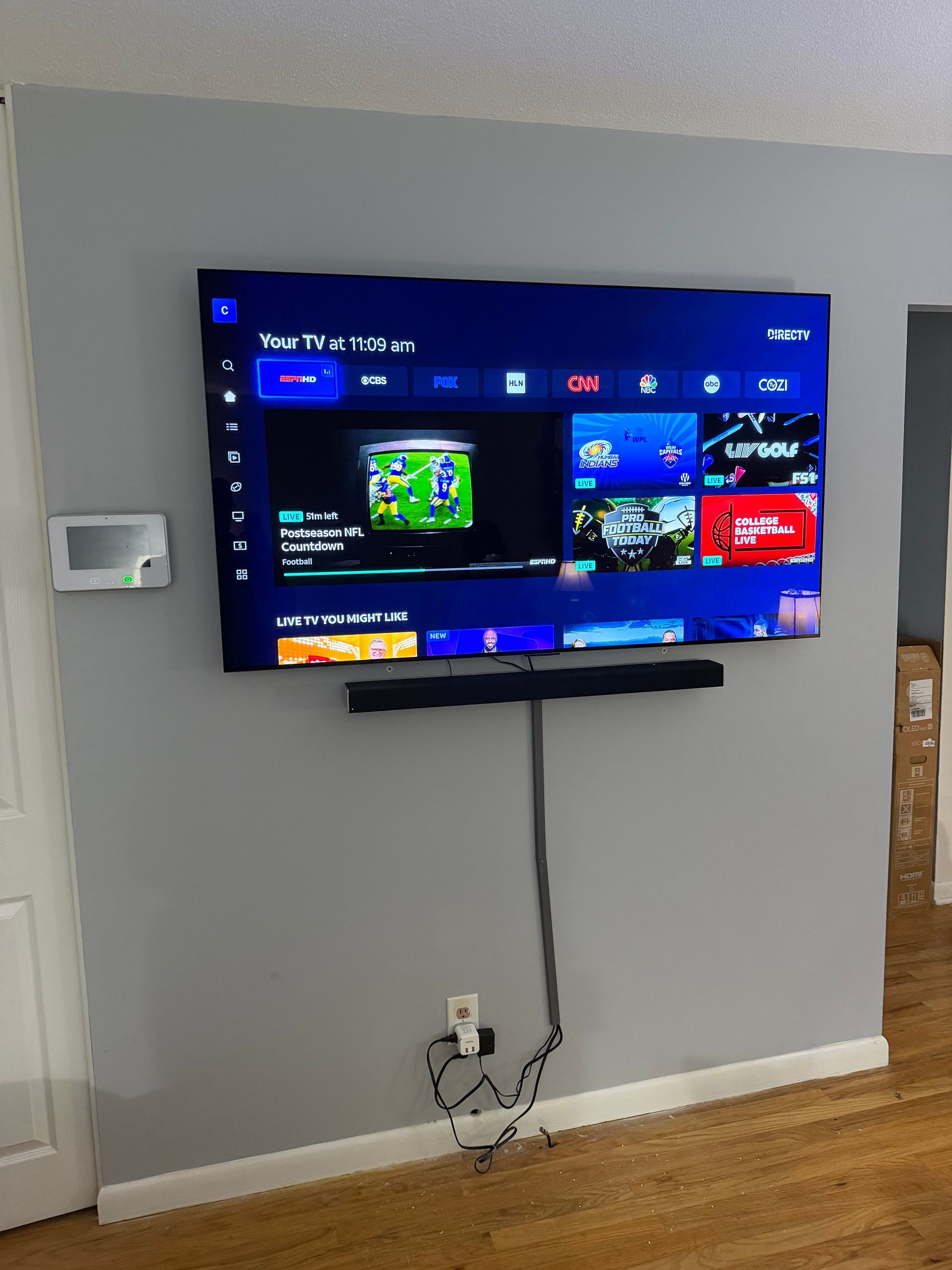 A large flat-screen TV mounted on a grey wall above a soundbar, with power cords hanging to a wall outlet below.