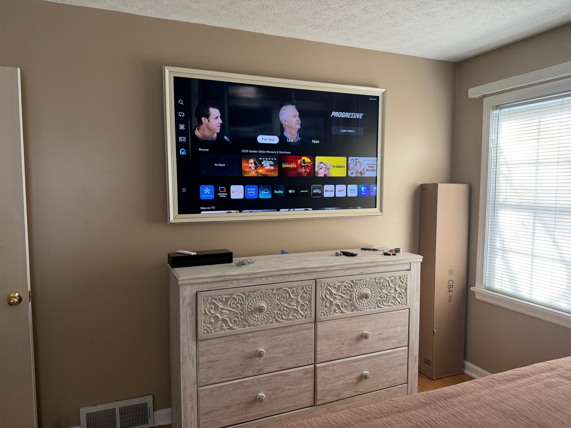A flat-screen TV in a decorative frame mounted above a weathered white wooden dresser in a bedroom with beige walls.