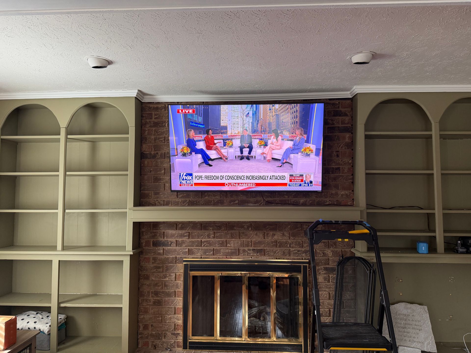 A television mounted on a brick chimney breast between two sets of built-in, arched bookshelves painted sage green.