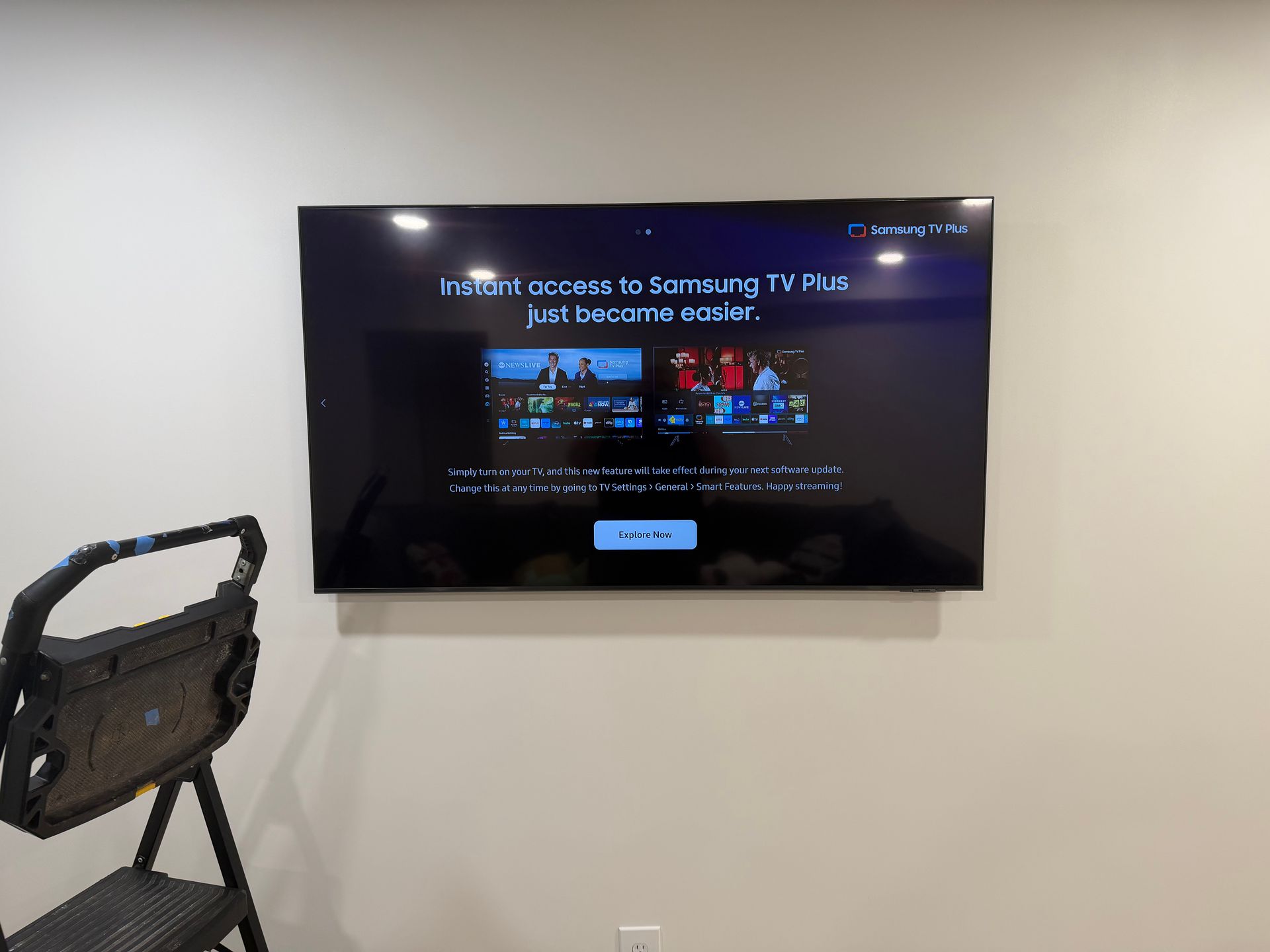 A mounted television screen displaying a Samsung TV Plus welcome prompt against a plain wall with a step stool nearby.