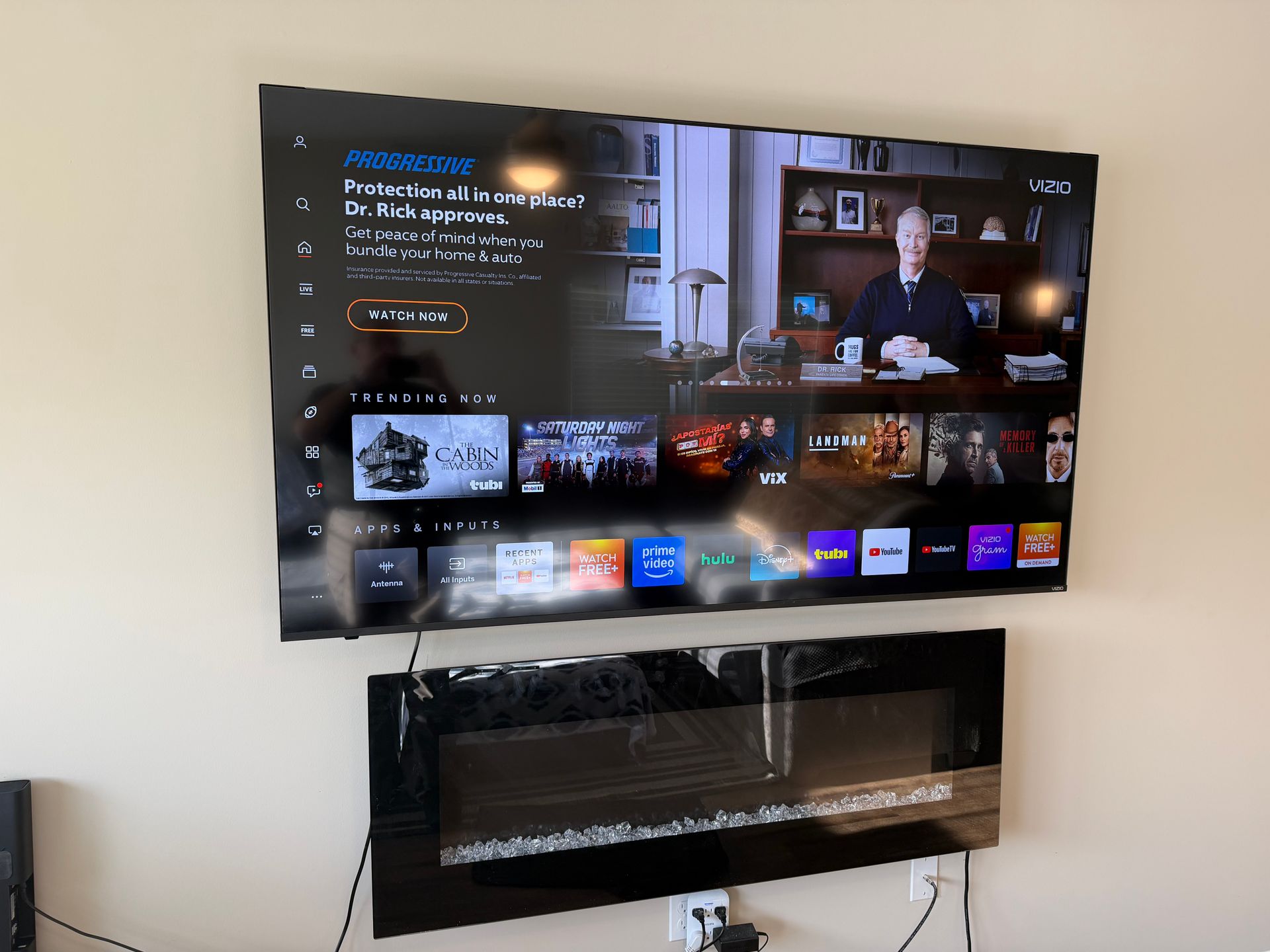 A large smart TV mounted on a wall above a black electric fireplace, displaying a streaming service interface.