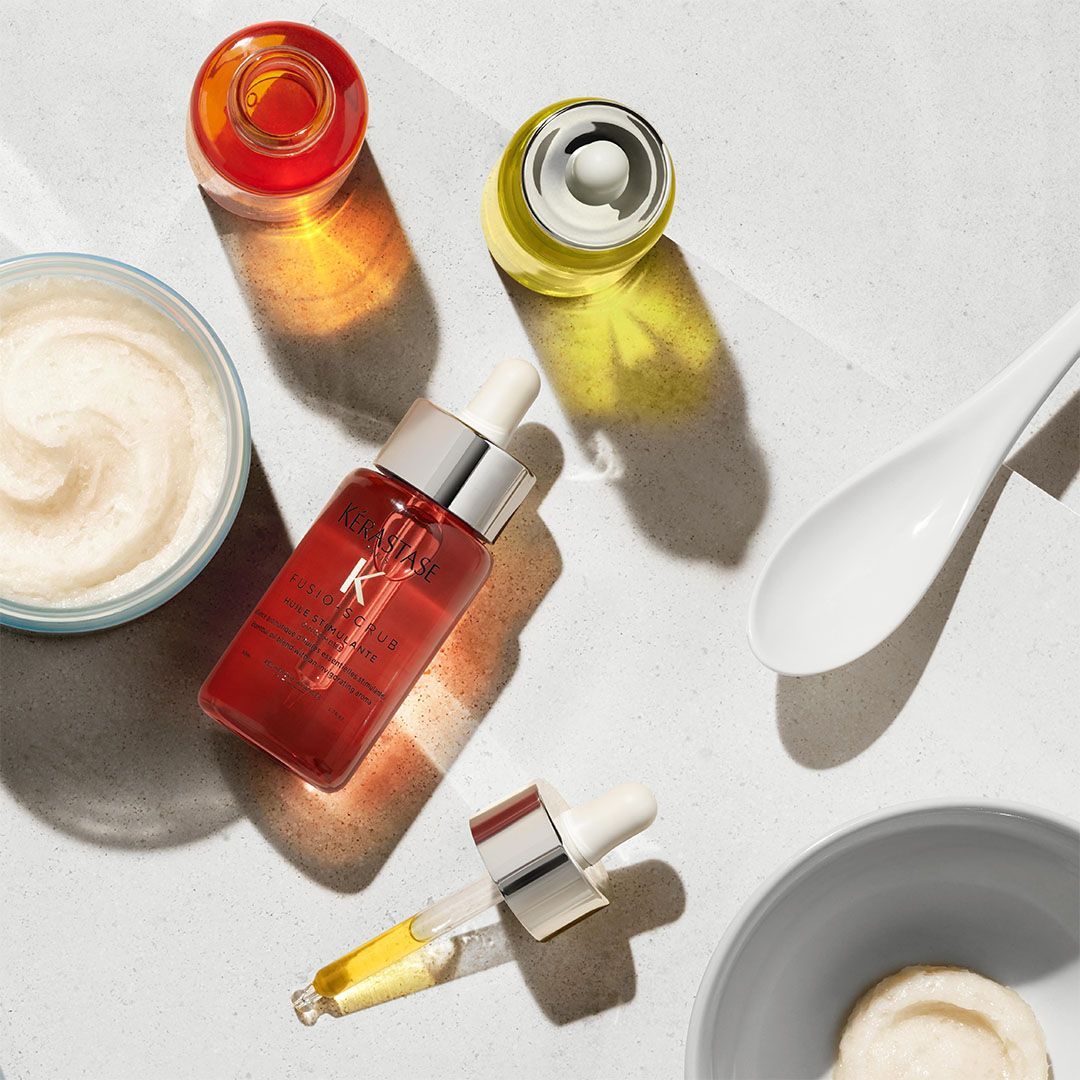 A bottle of serum is on a table next to a bowl of cream and a spoon