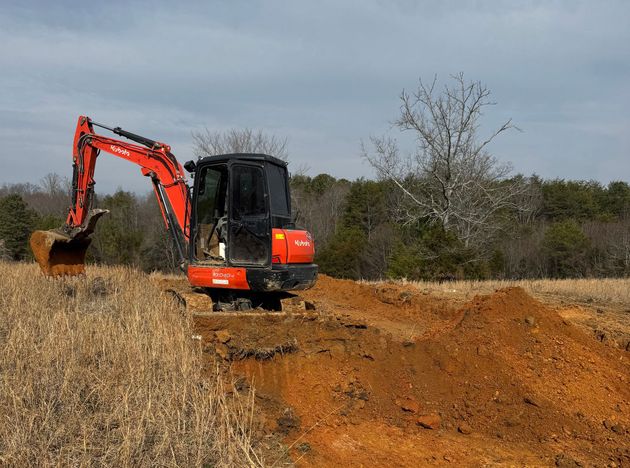 An orange excavator digs into red soil in a grassy field with trees in the background under a cloudy sky.
