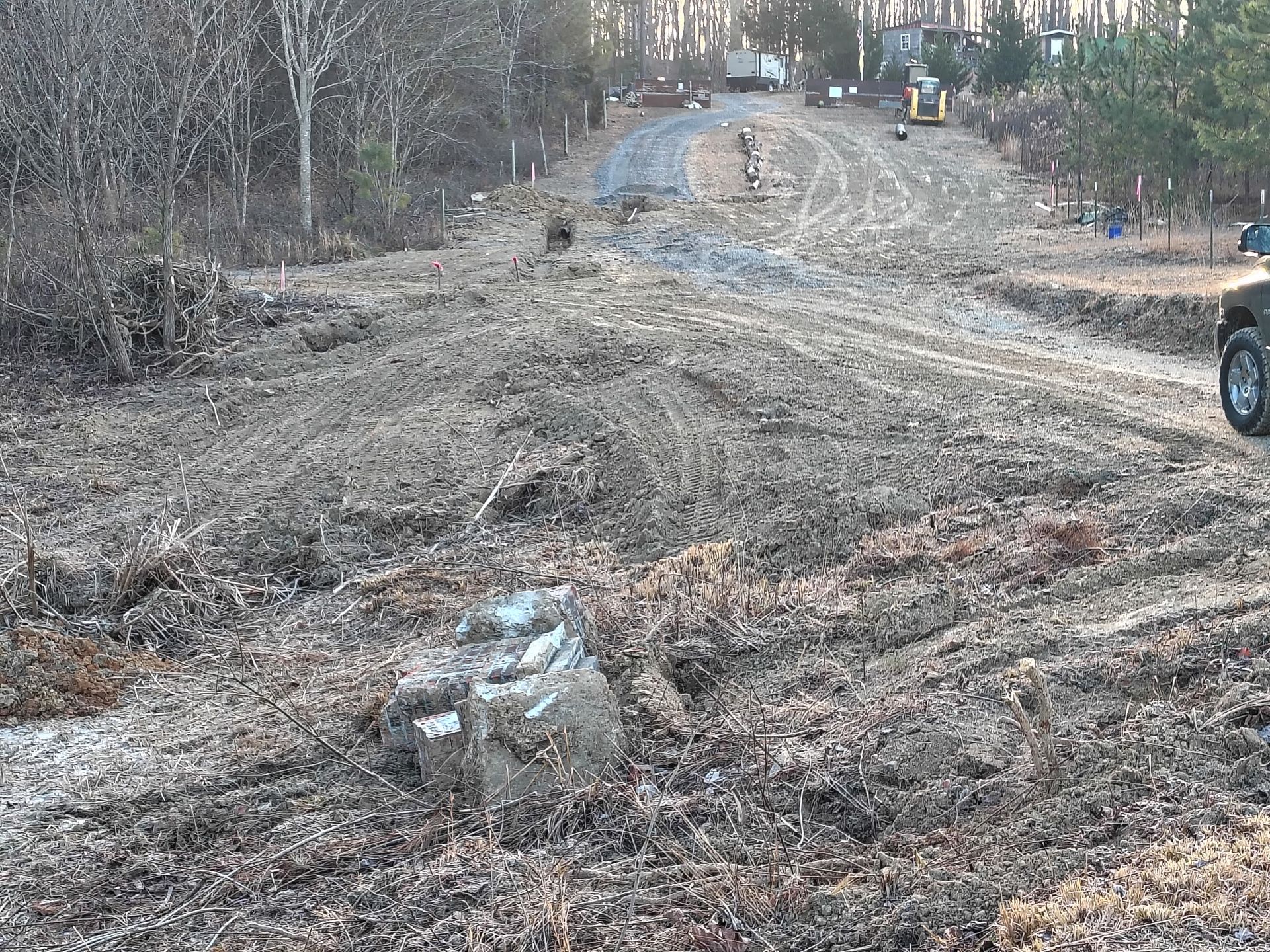 A construction site with a freshly cleared dirt path and a gravel driveway leading toward a vehicle in a wooded area.