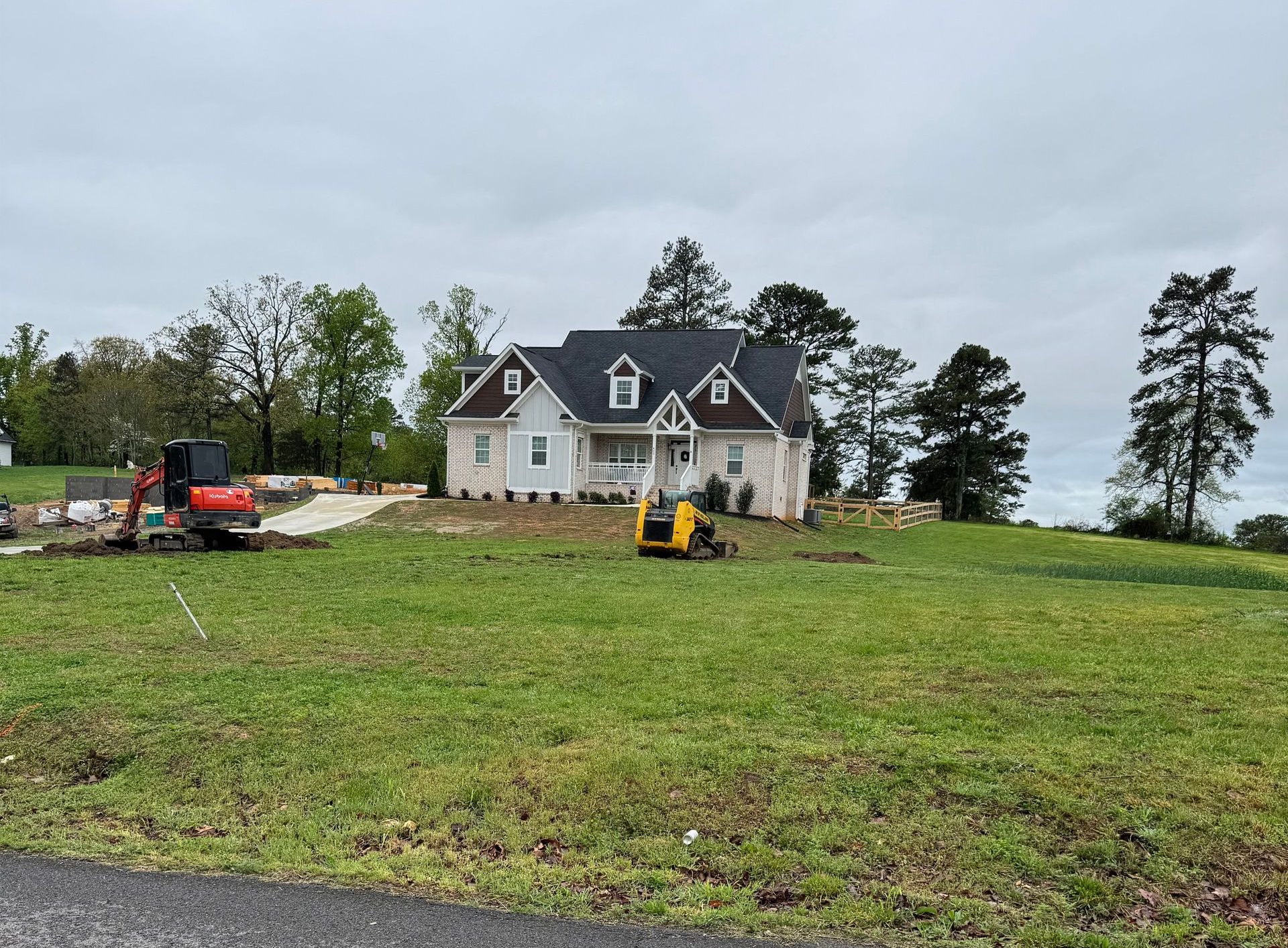 A white house with a dark roof sits on a grassy hill, with a small excavator and a skid steer parked in front.