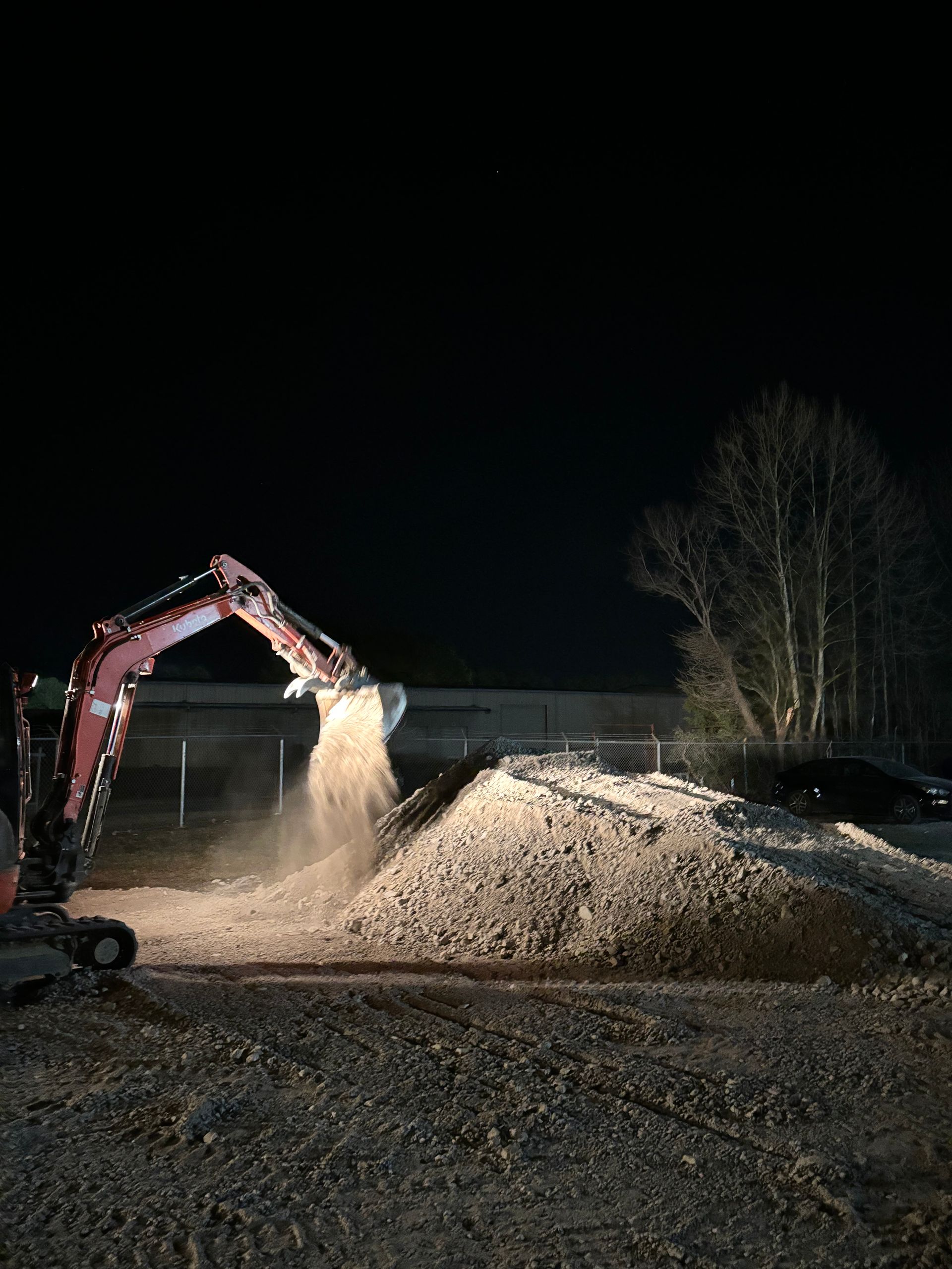 A red excavator pouring sand onto a large pile at night under artificial lighting.