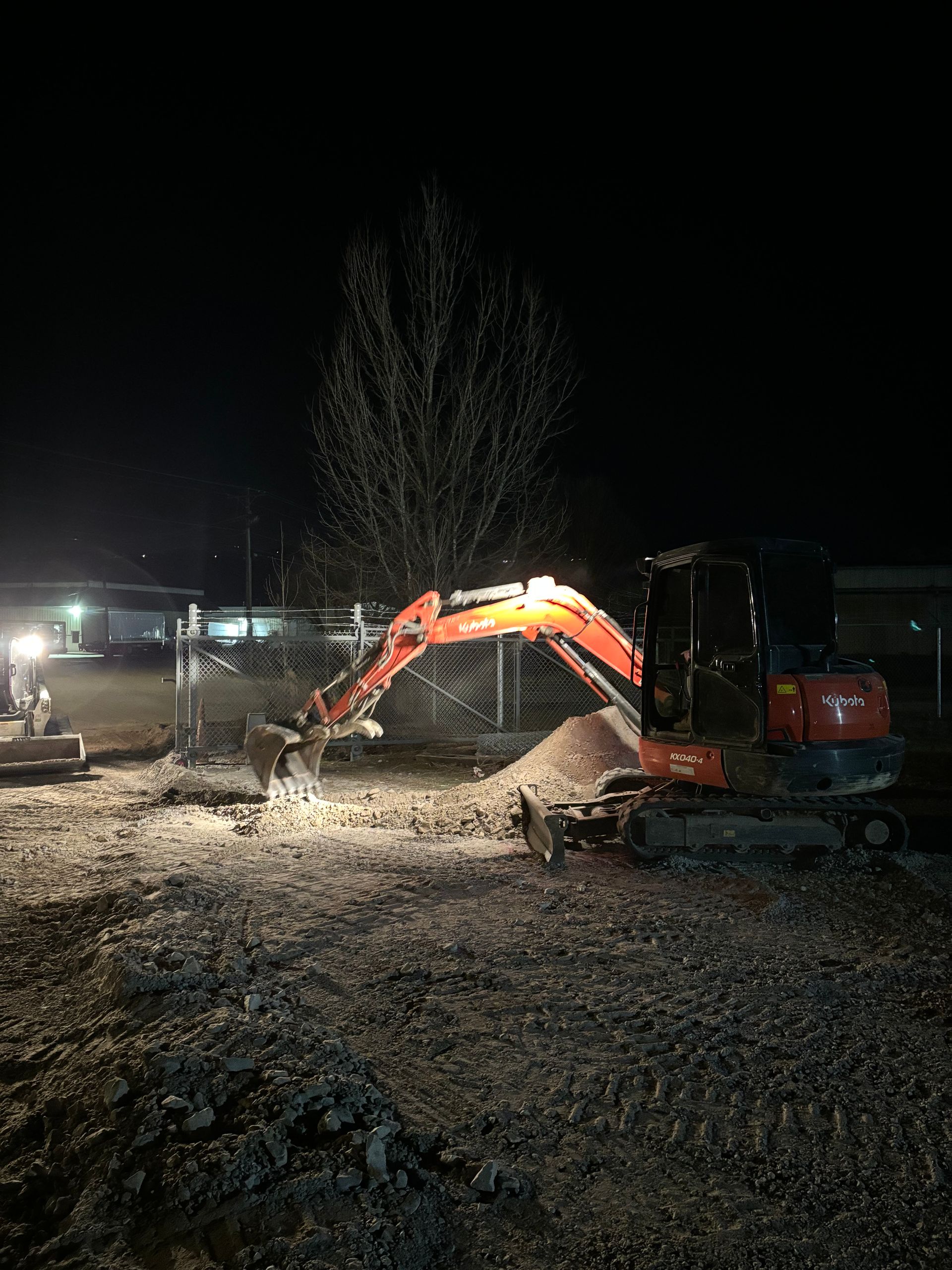 An orange excavator works on a gravel construction site at night under bright work lights.