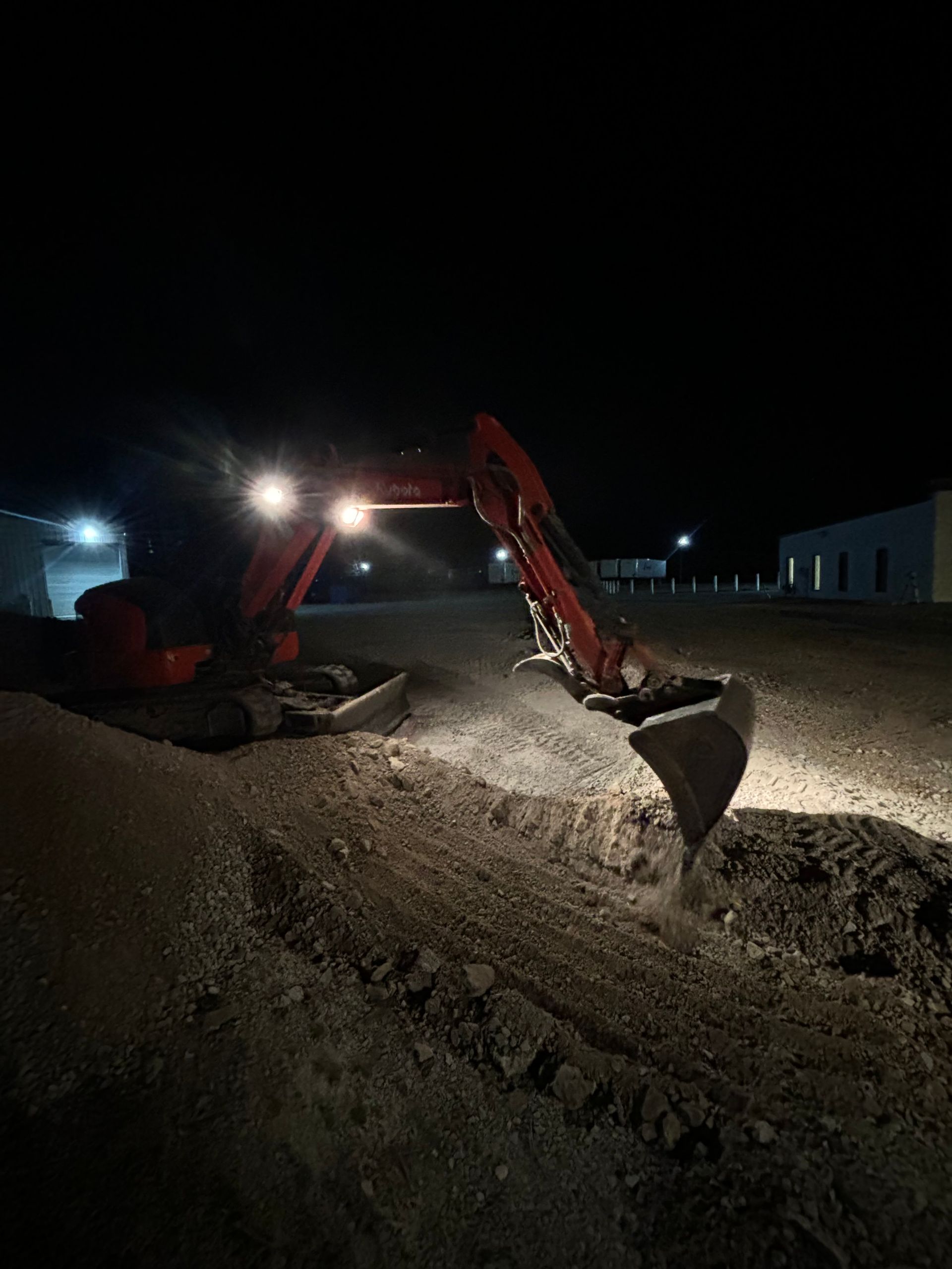 A red excavator with bright lights digging into gravel on a construction site at night.