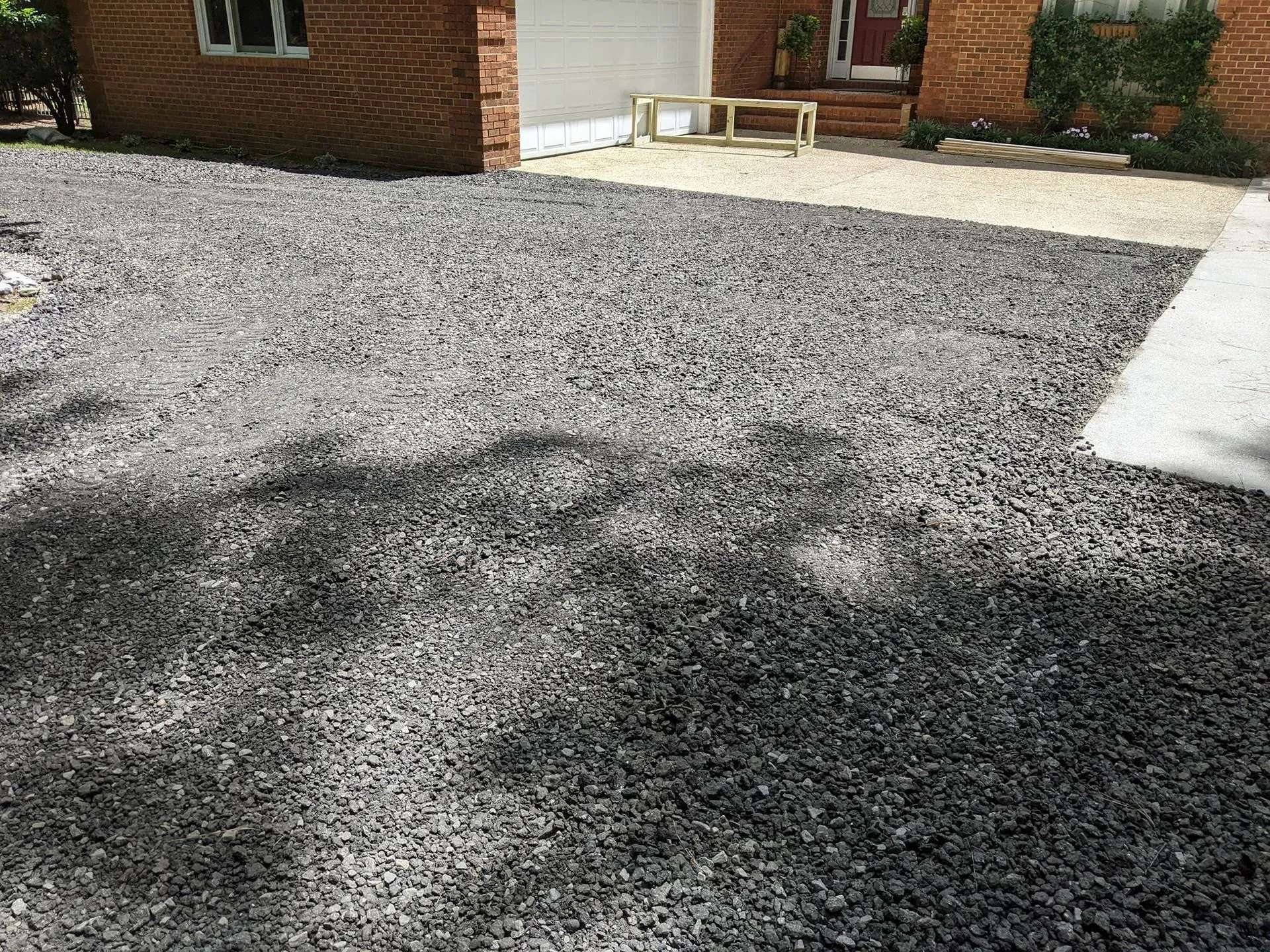 Gravel driveway beside a brick house, with a bench and leafy shadows in the sunlight.
