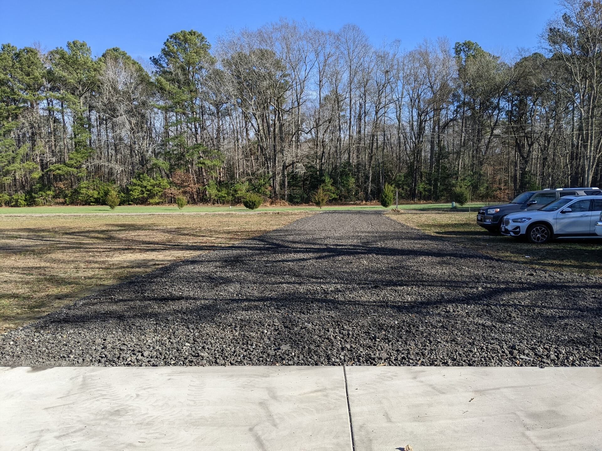 Gravel parking lot beside a wooded tree line, with several cars parked on the right.