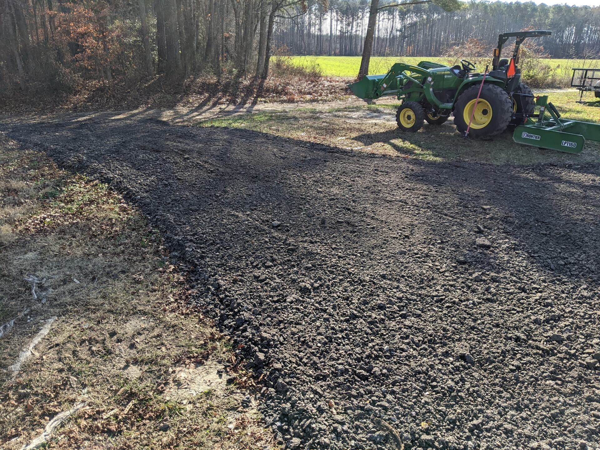 Tractor mowing a gravel path beside a wooded field on a sunny day.