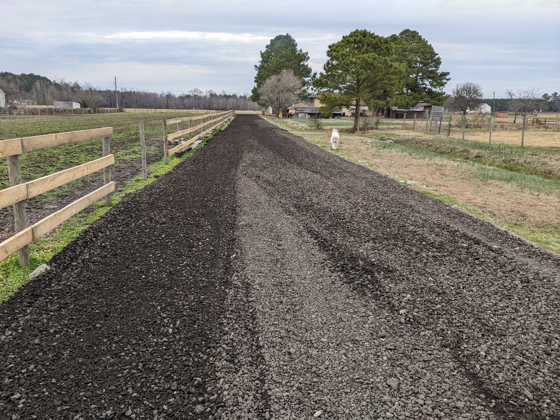 Freshly leveled dark gravel road stretching through rural fields under a cloudy sky