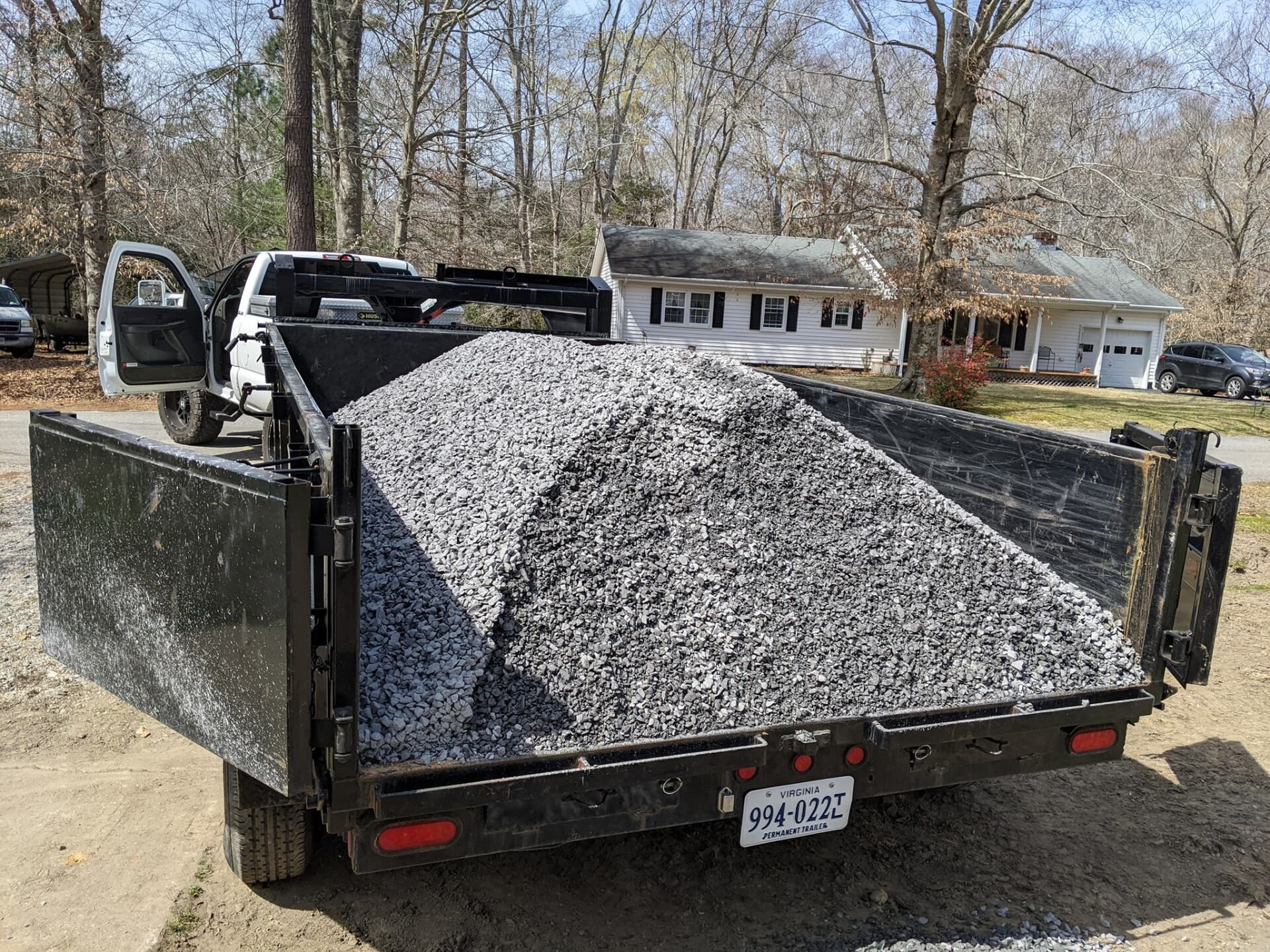 Dump truck bed filled with gray gravel in a driveway, with houses and bare trees in the background.