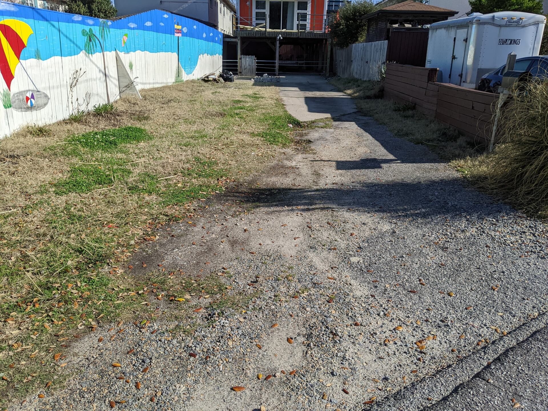 Gravel driveway beside a grassy yard and colorful mural fence leading to a house and shed.