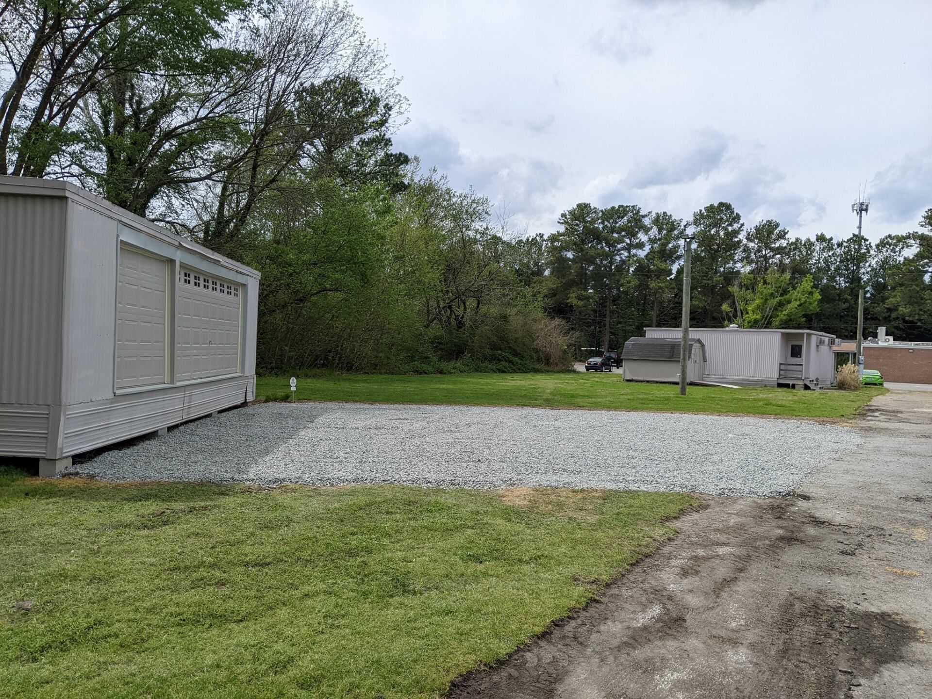 Gravel lot with two white portable storage containers beside trees and grass under a cloudy sky