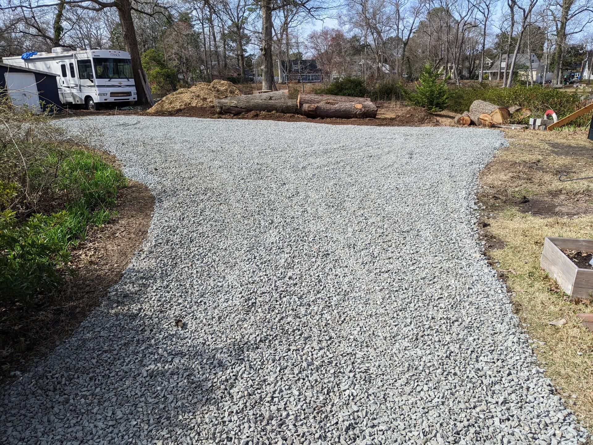 Gravel driveway leading to a wooded yard, with an RV parked on the left and rocks along the back edge