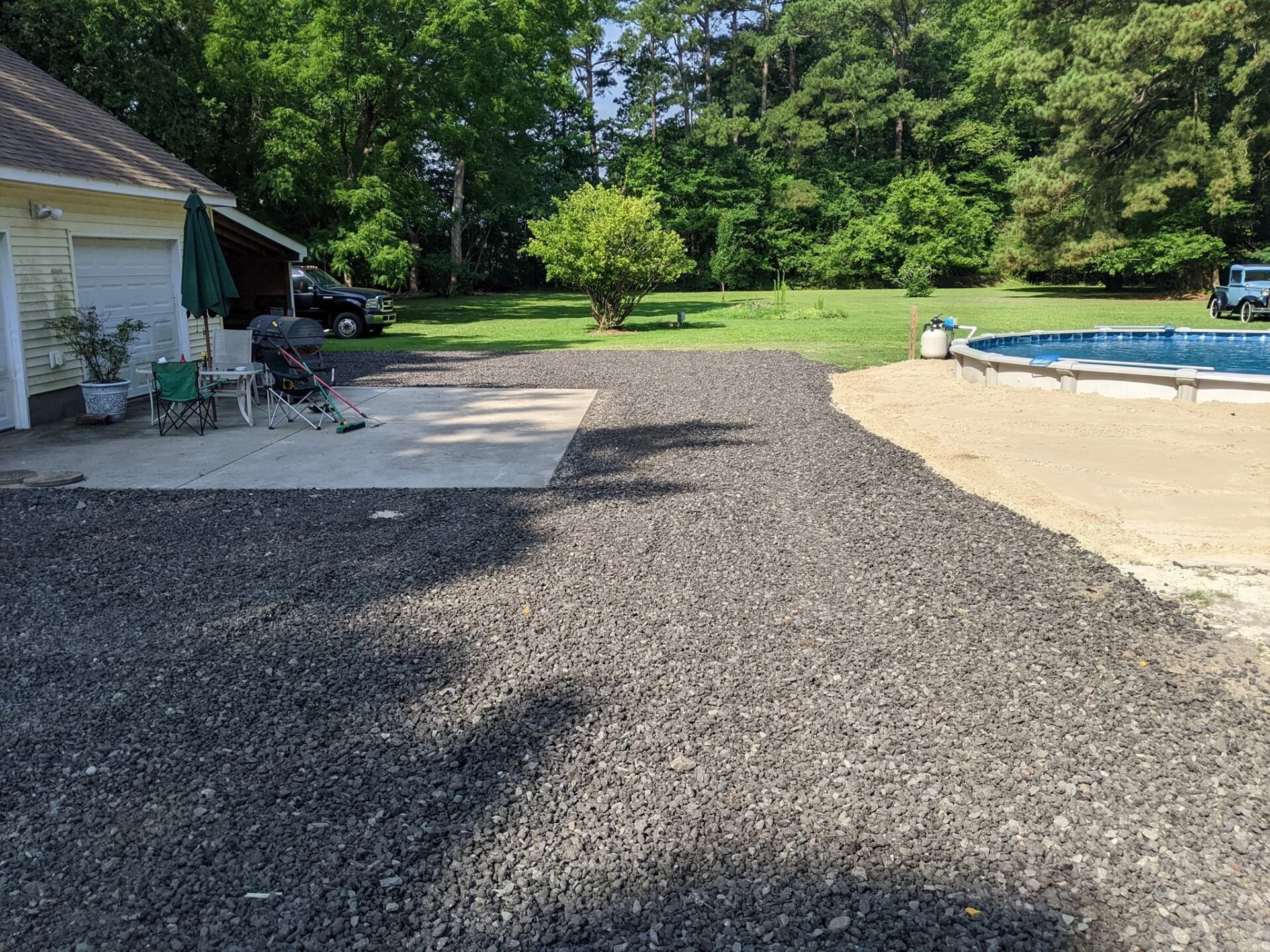 Gravel driveway beside a house and patio, leading to a backyard with a swimming pool and trees.