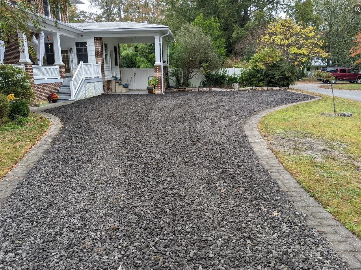 Gravel driveway leading to a white house with a porch, bordered by grass and trees.