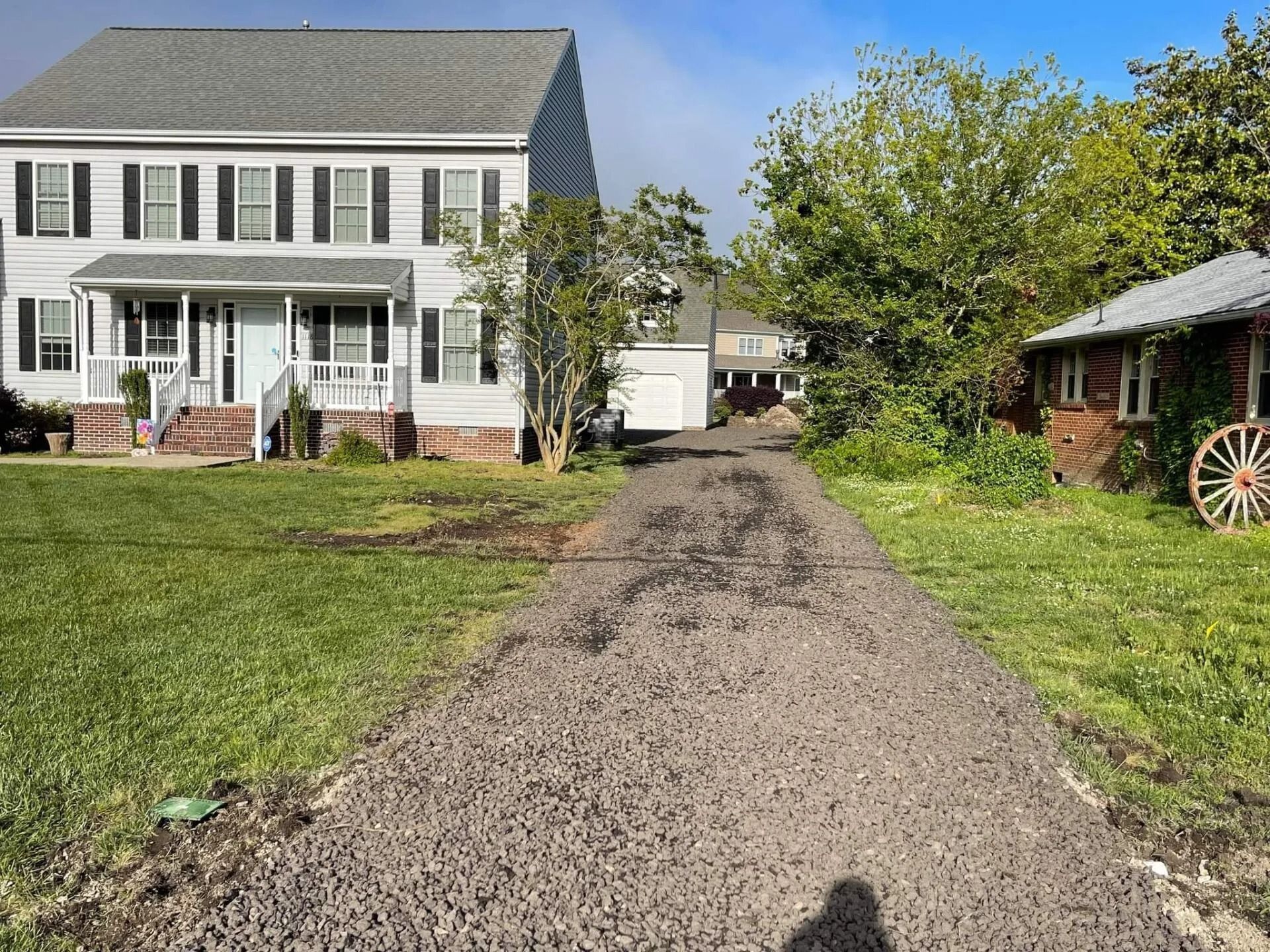 Gravel driveway leading to a white farmhouse with a porch, green lawn, and trees on a sunny day