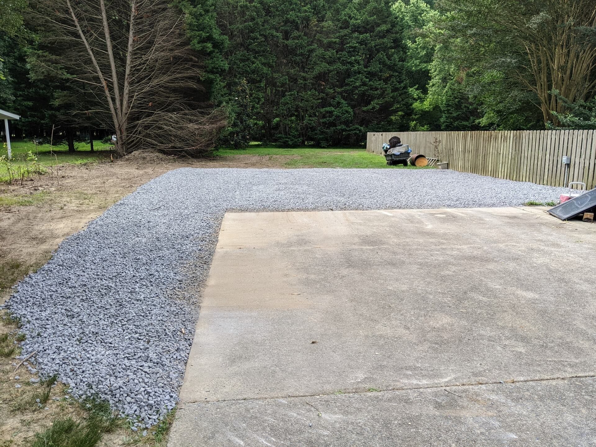 Gravel driveway beside a concrete pad, with a wooded yard and a fence in the background.
