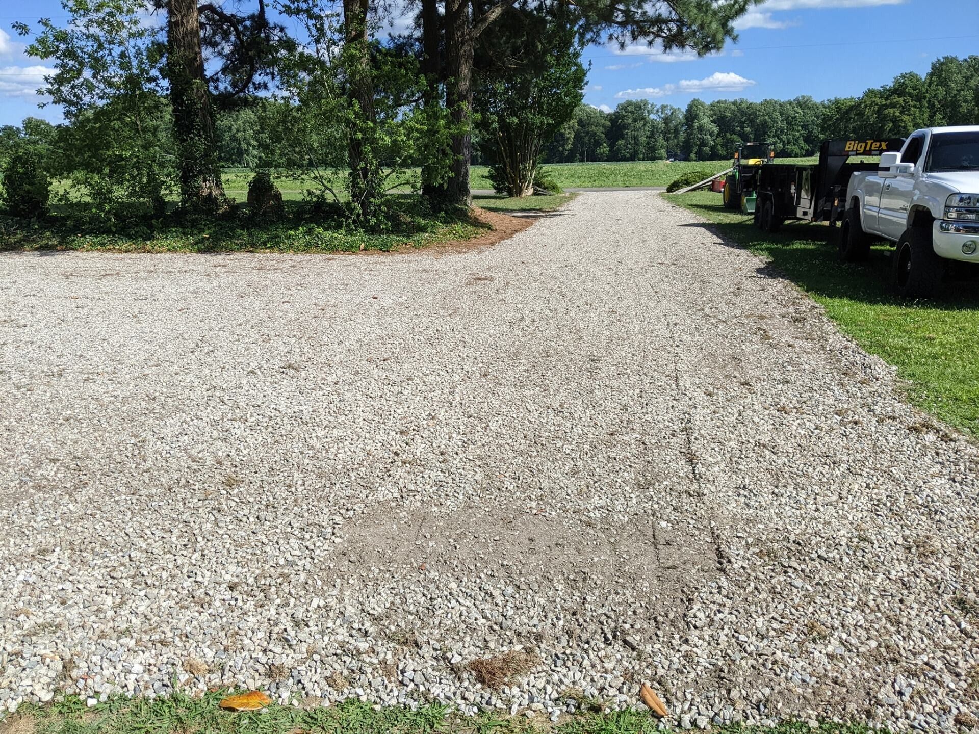 Gravel driveway lined with trees, leading past parked vehicles on a sunny day