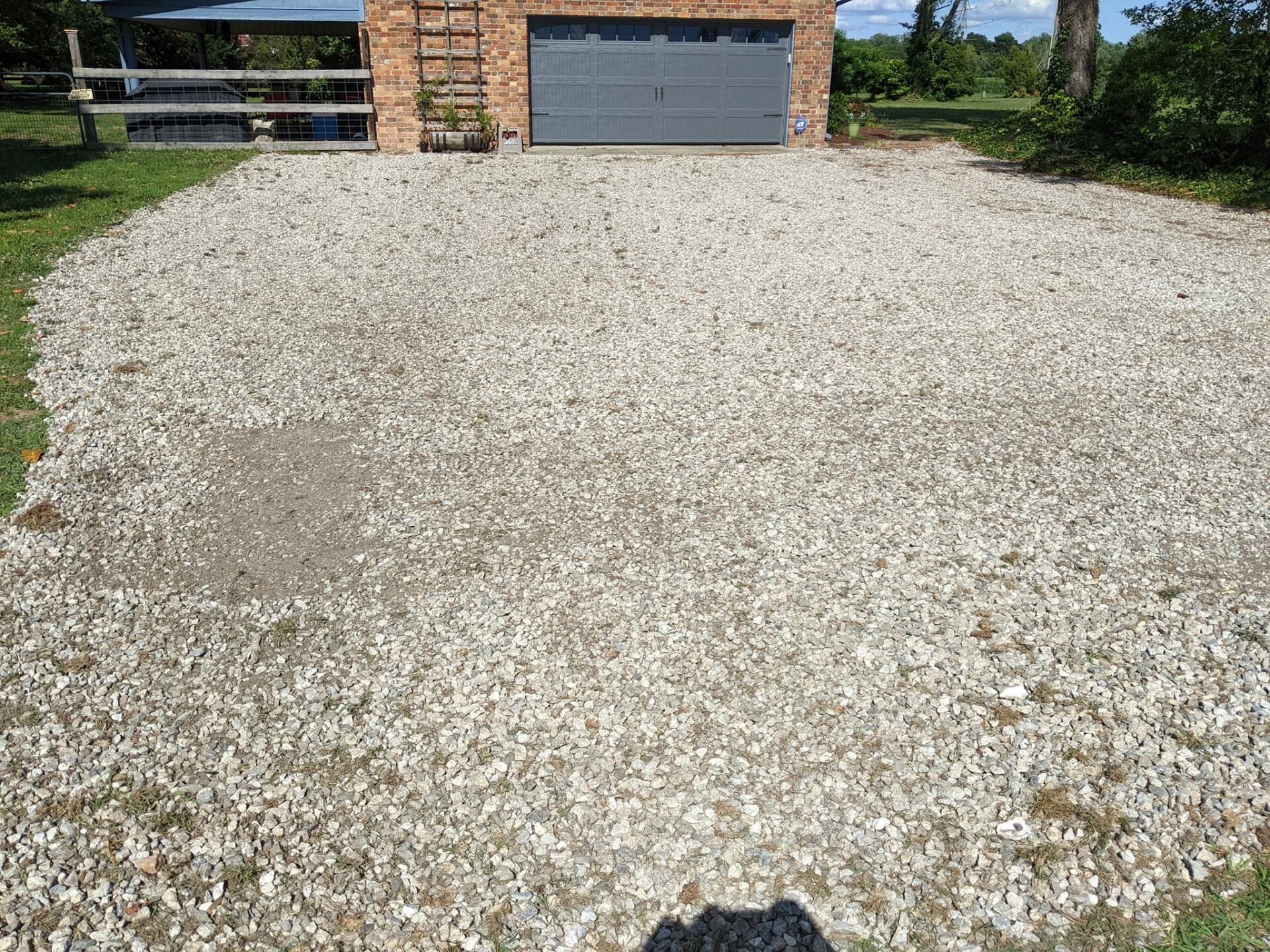 Gravel driveway leading to a brick garage with a closed gray door, bordered by grass and trees