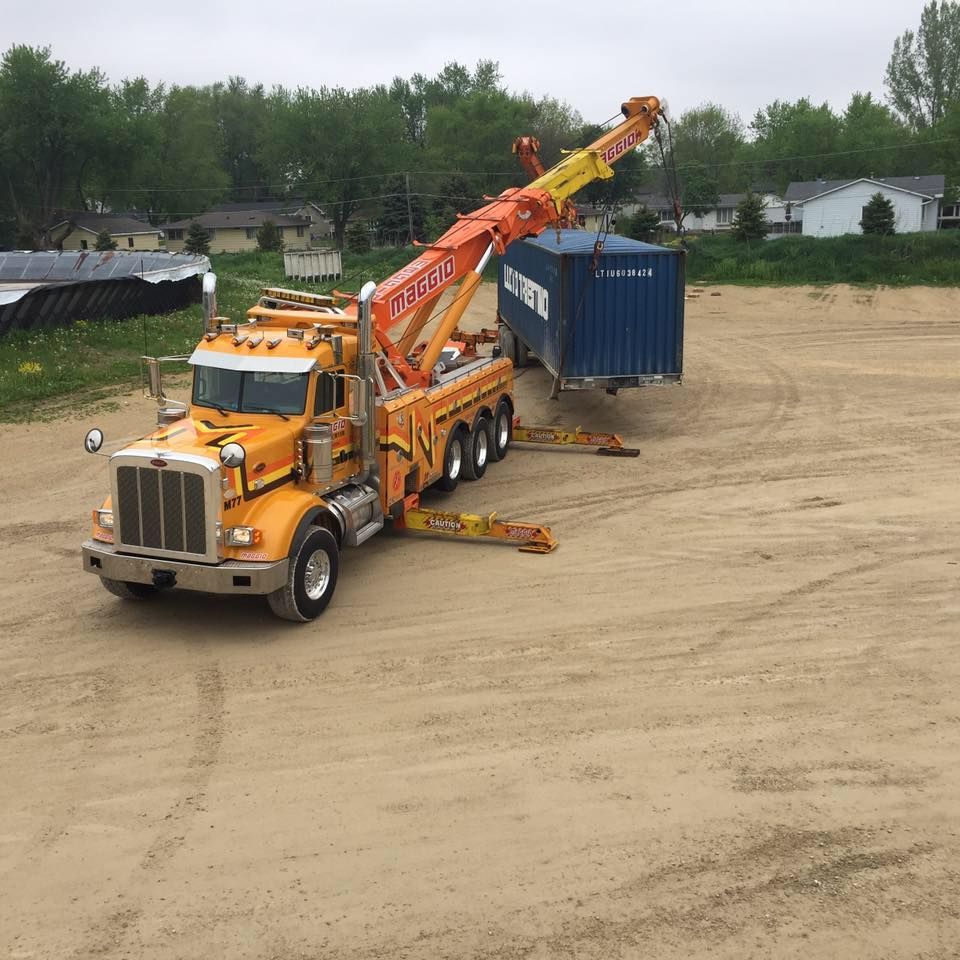 Yellow tow truck lifting a blue shipping container on a dirt lot.
