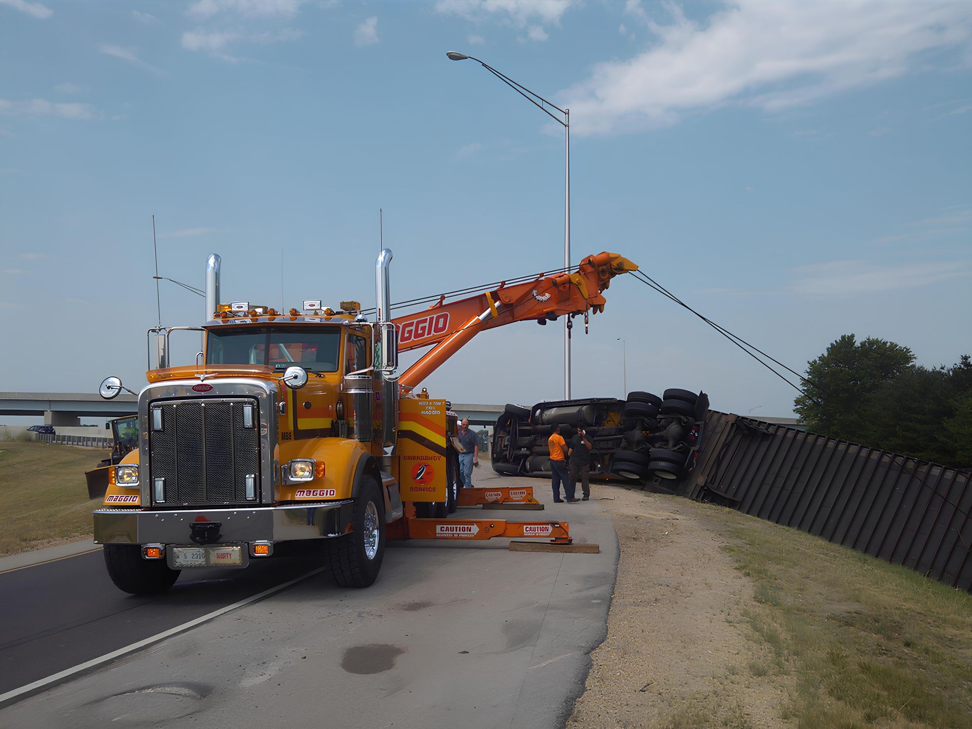Yellow tow truck lifting a tipped-over black semi-truck on a roadside. Two people in orange vests.