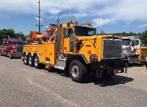 Yellow heavy-duty tow truck with crane, parked on pavement, alongside other tow trucks. Blue sky in the background.