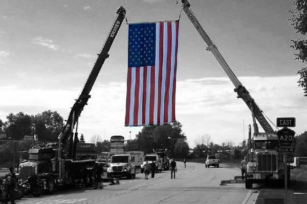 Two cranes suspending a large American flag over a road; black and white scene with colored flag.