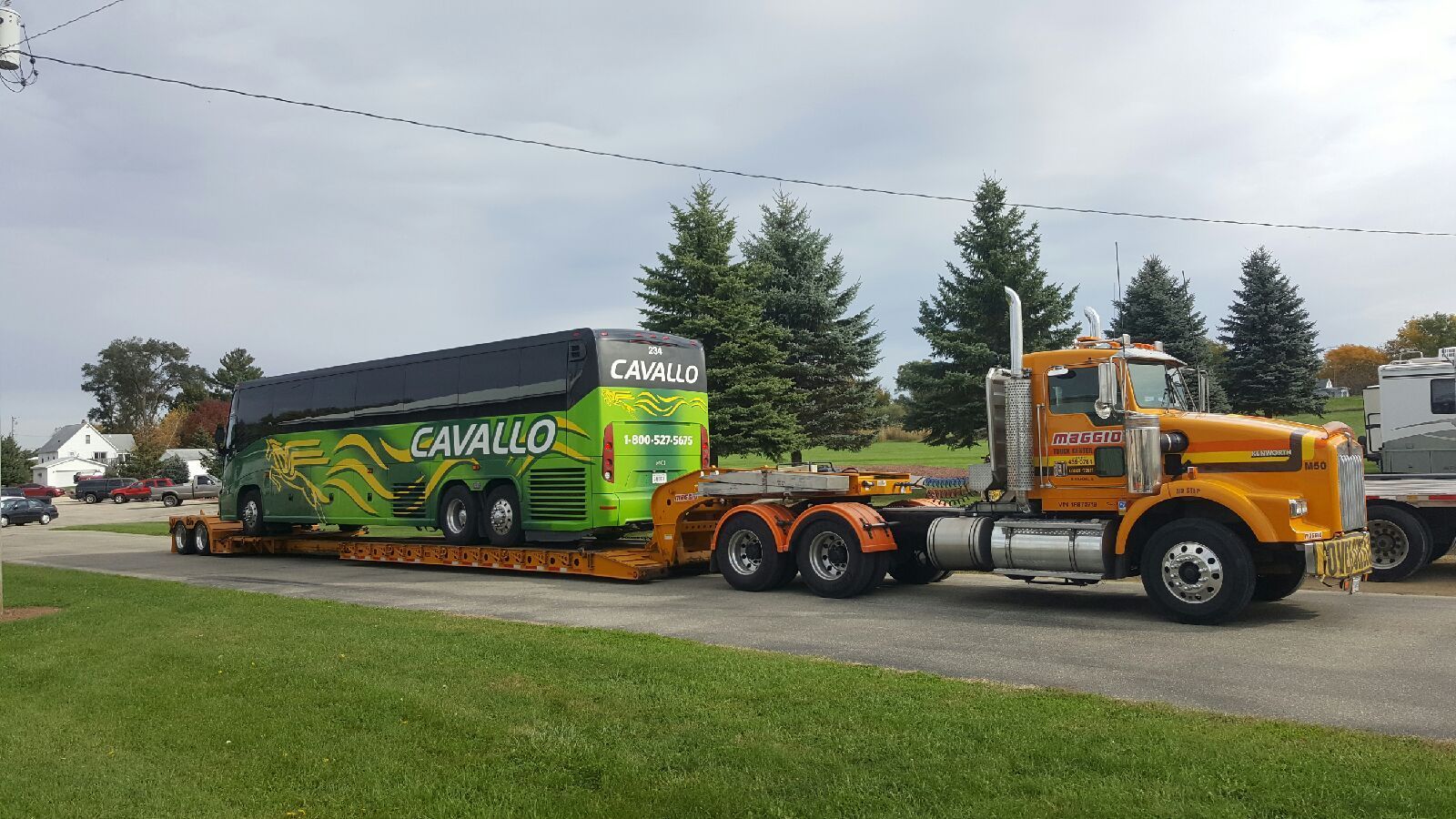 Yellow semi-truck towing a large green and black bus on a flatbed trailer, parked on a grassy area.