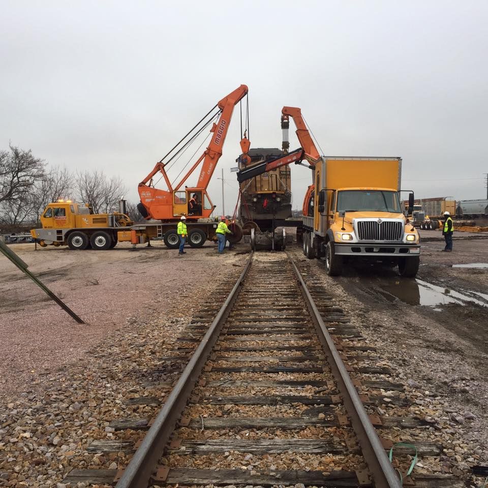 Two cranes lifting equipment from railcar onto truck bed on train tracks. Yellow trucks and workers.