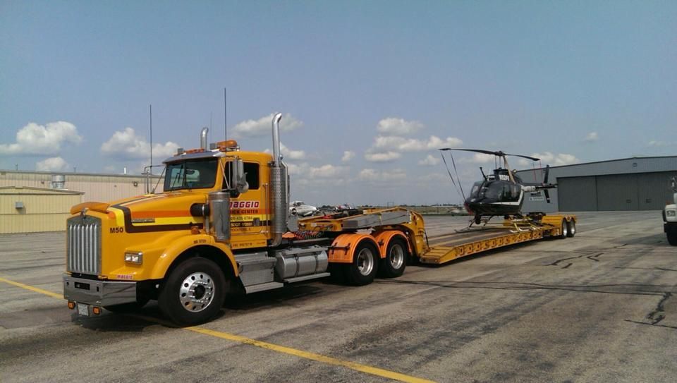 Yellow semi-truck transporting a helicopter on a lowboy trailer, outdoors.