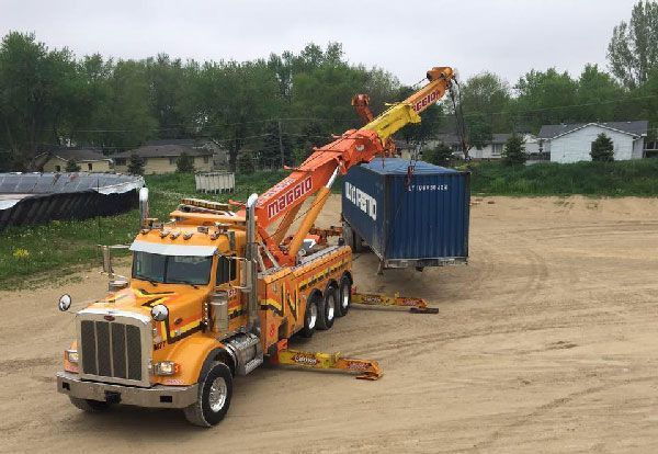 Orange tow truck lifting a blue shipping container on a dirt lot.