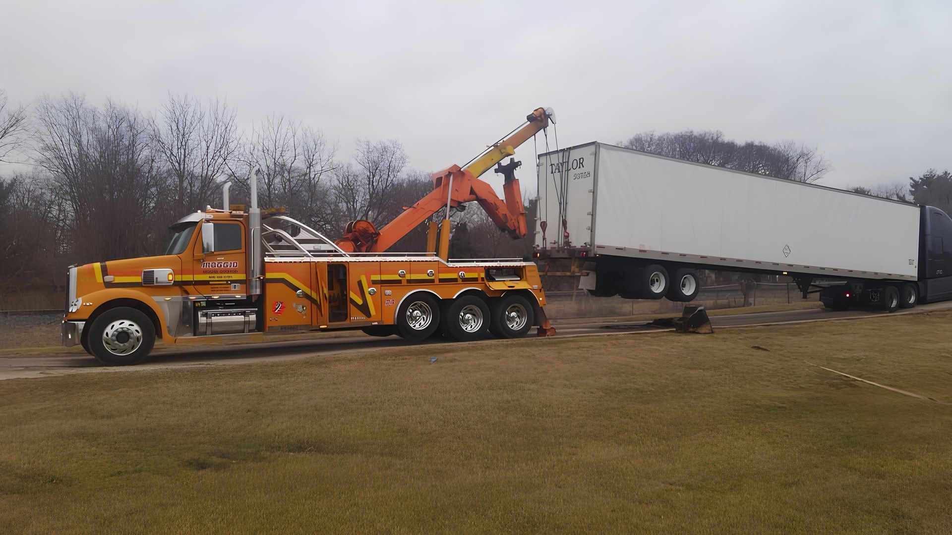 Orange tow truck lifting a white semi-trailer on a cloudy day.
