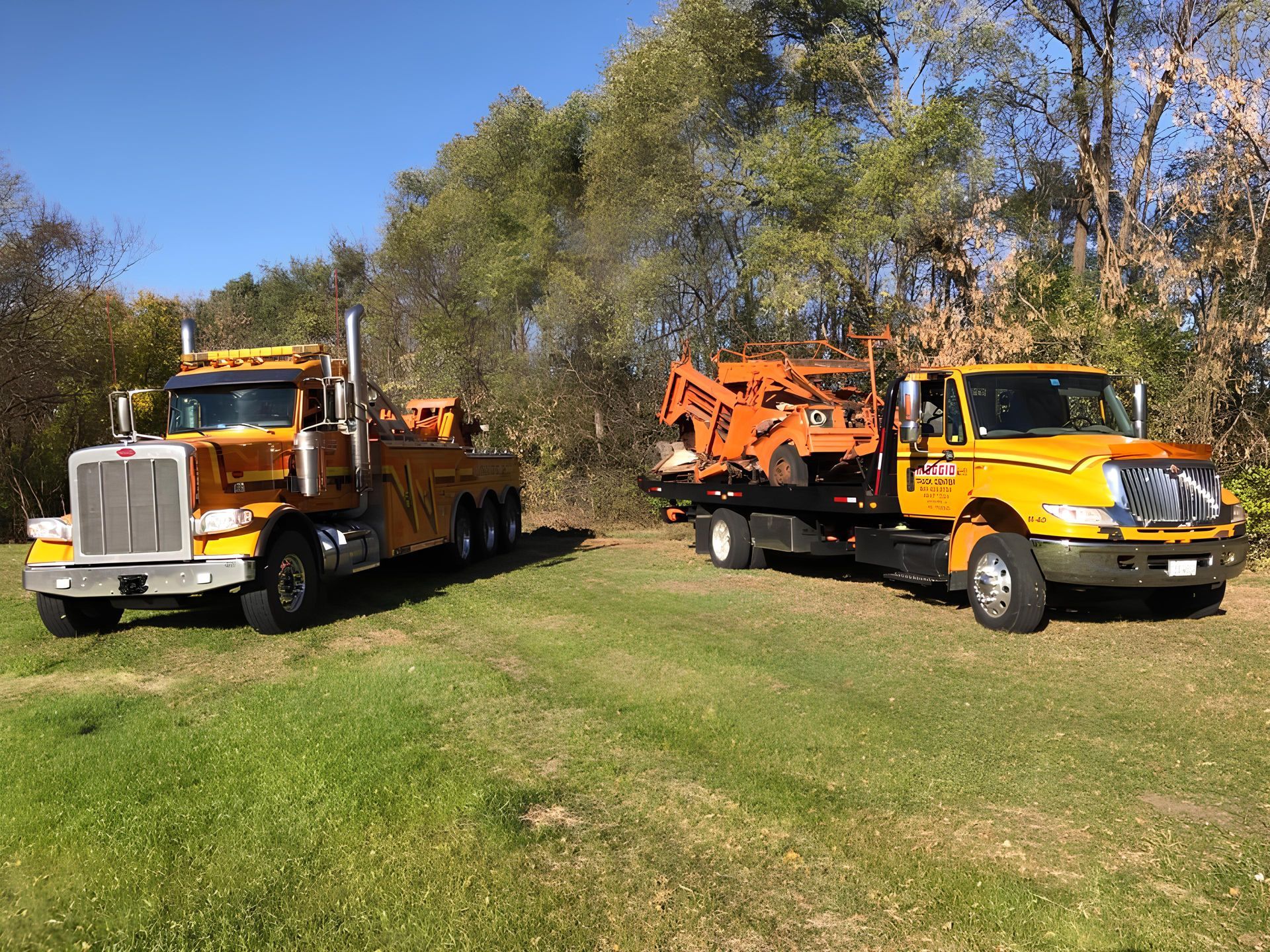 Two yellow tow trucks on grass, one hauling an orange front loader, trees in the background under a blue sky.