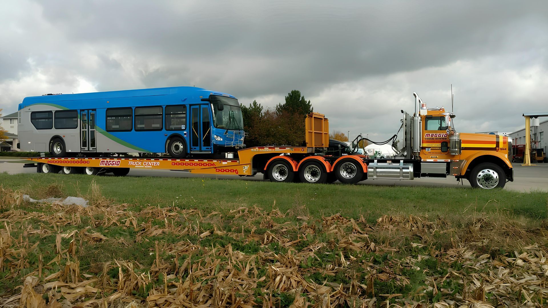 A large orange heavy duty truck hauling a blue transit bus on a flatbed trailer, in an outdoor setting.