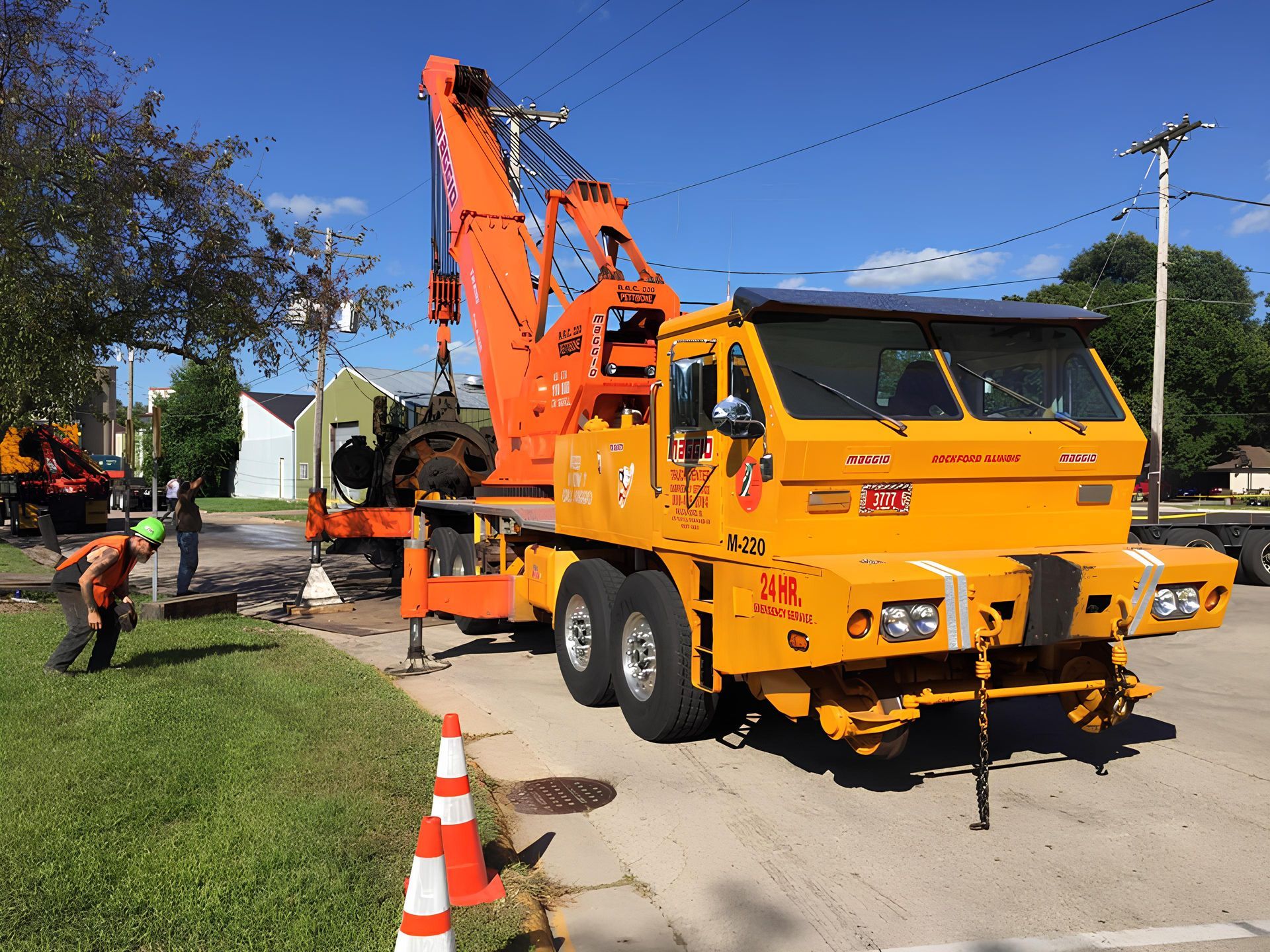 Orange crane truck lifting equipment; workers in safety vests; street setting with homes and grass.