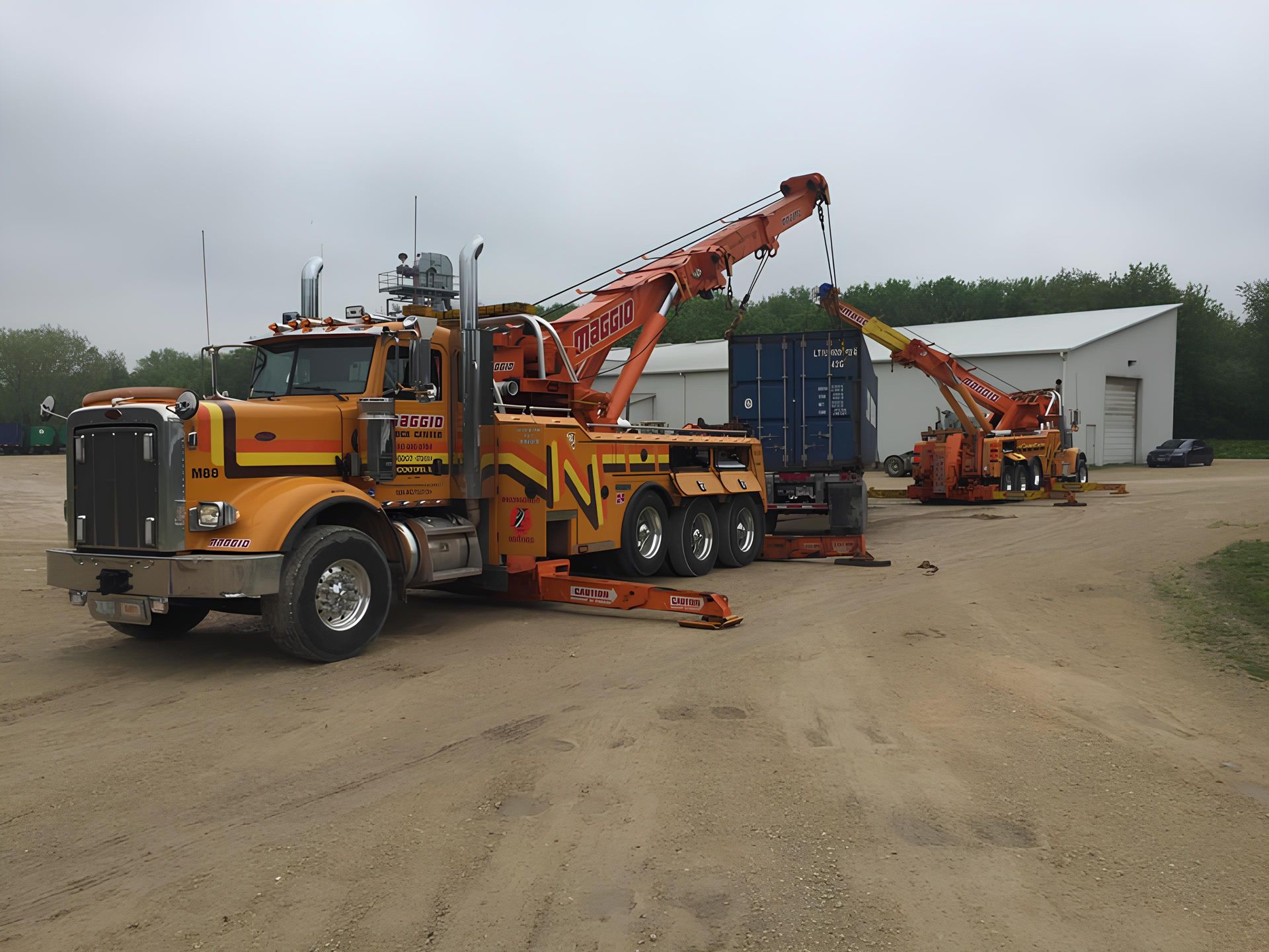 Two orange heavy duty tow trucks lifting a blue cargo container on a gravel lot near a white building.
