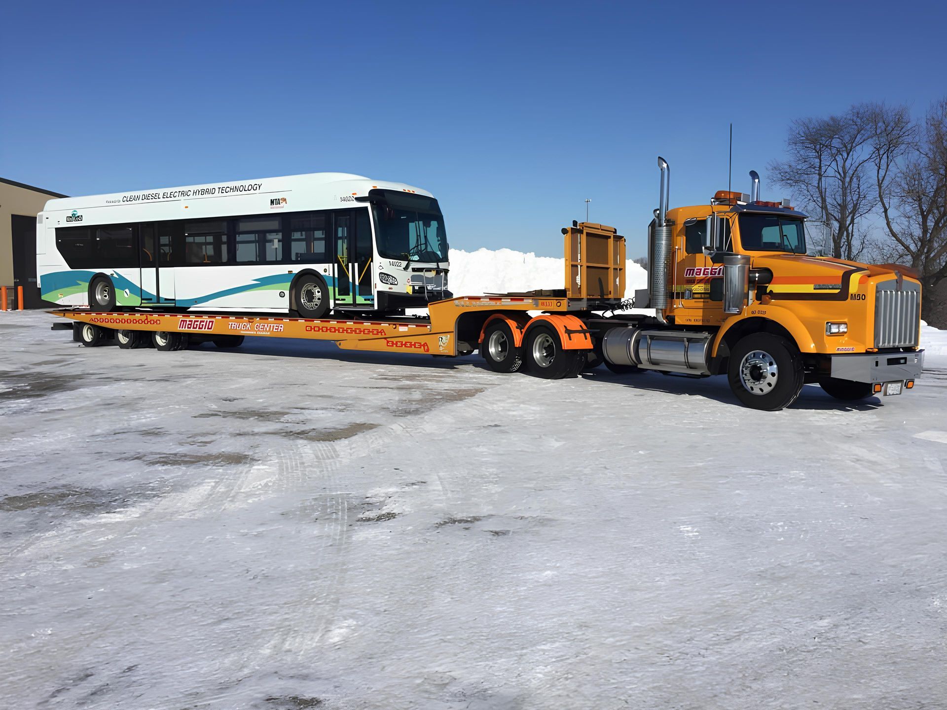 Yellow semi-truck transporting a city bus on a flatbed trailer across snow-covered ground under a clear blue sky.