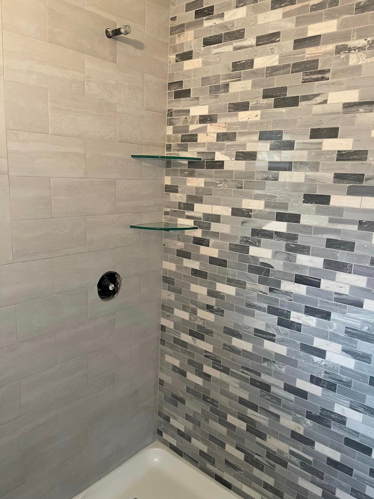A bathroom with a tiled wall , tub , shower and shelves.