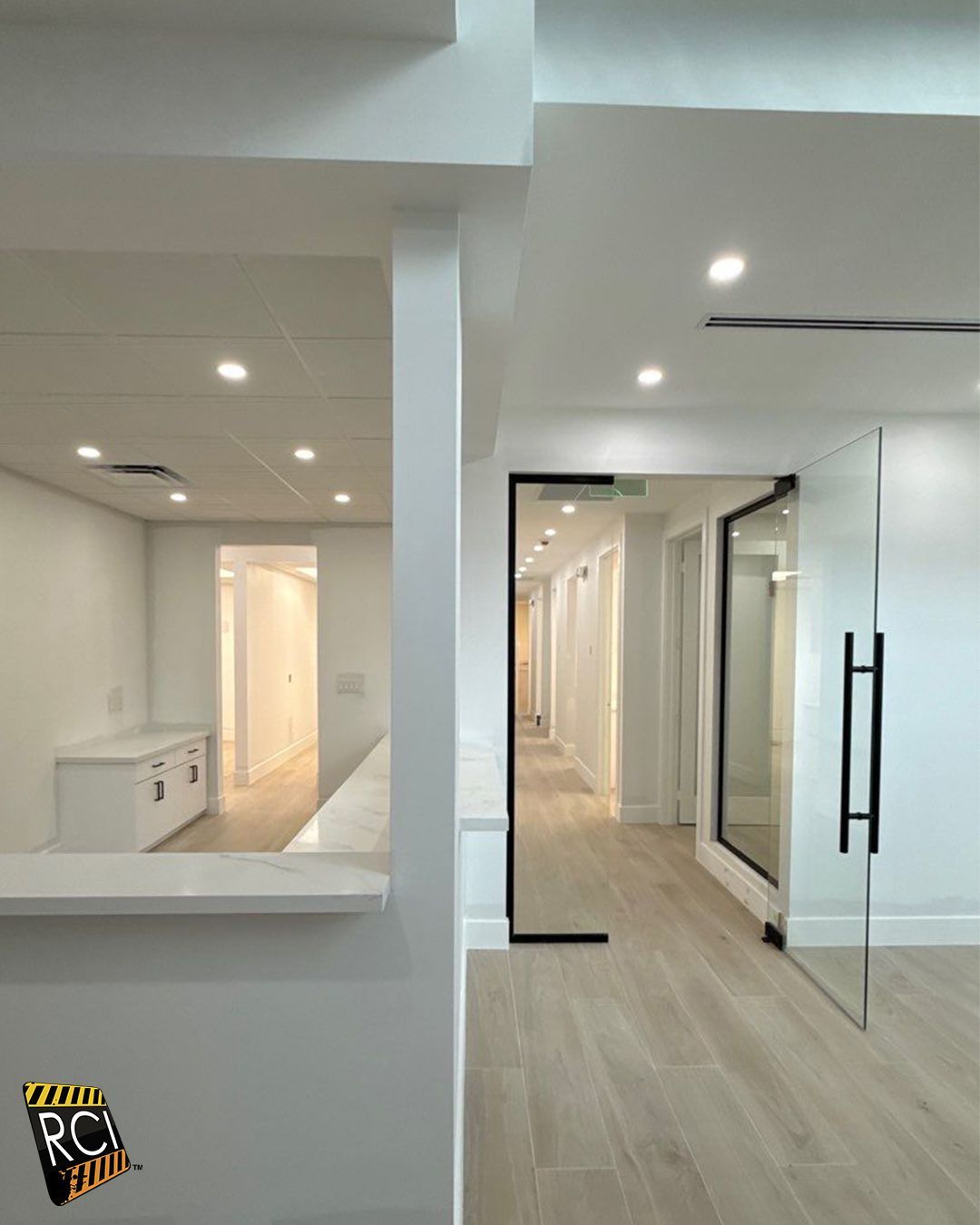 Interior view of a white office space with a hallway, glass door, and light wood flooring.